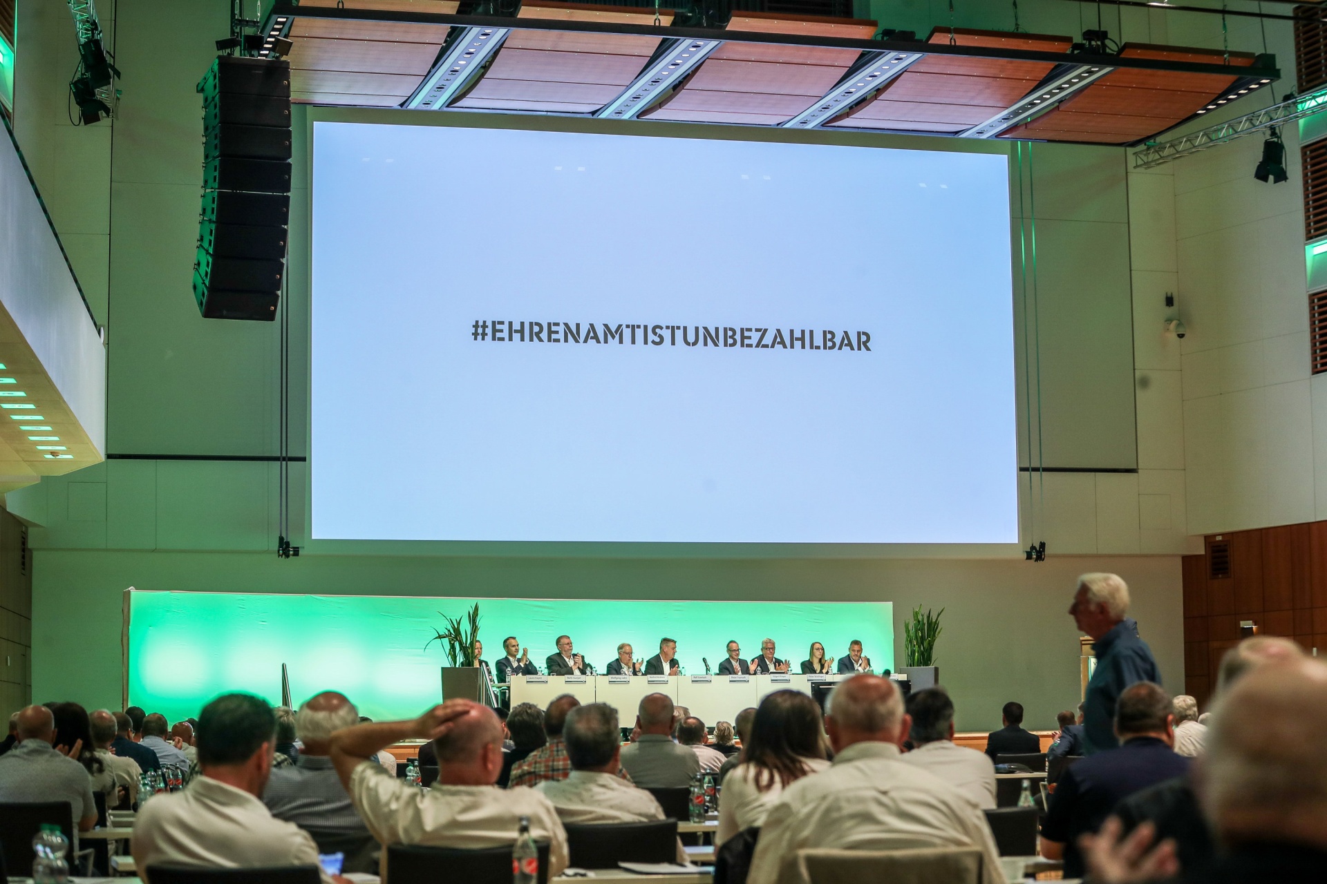 [{"clang_id":1,"value":"Großer Konferenzraum mit Bühne. Auf der Leinwand \"#EHRENAMTISTUNBEZAHLBAR\". Publikum sitzt vor einer Reihe von Personen am Tisch."}]