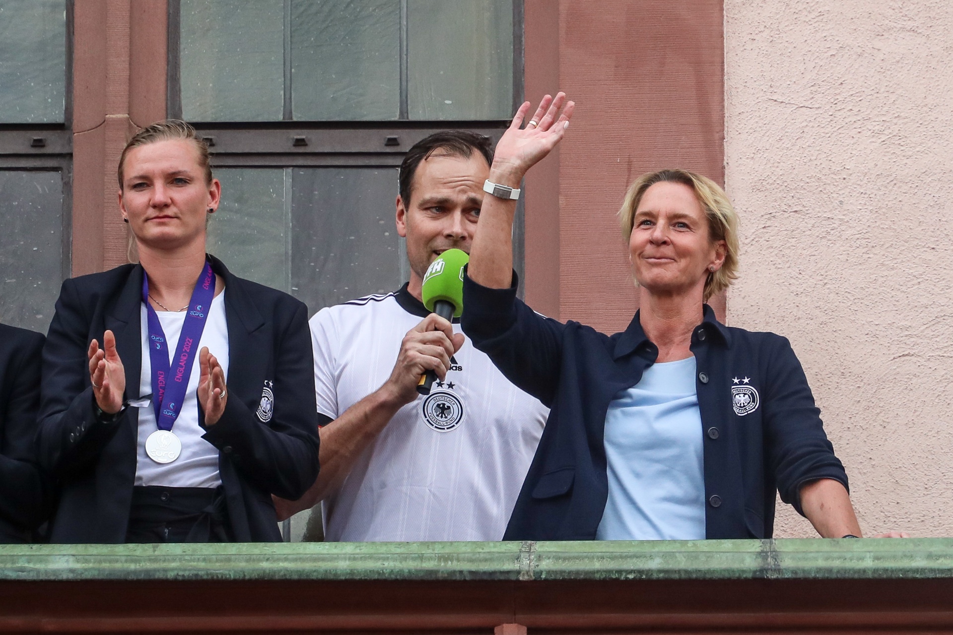 [{"clang_id":1,"value":"Drei Personen stehen auf einer erhöhten Plattform vor einem Gebäude. Links eine Frau im schwarzen Blazer, in der Mitte ein Mann mit Mikrofon und weiß-schwarzem Trikot, rechts eine Frau im blauen Hemd und schwarzem Blazer. Alle drei lächeln und winken."}]