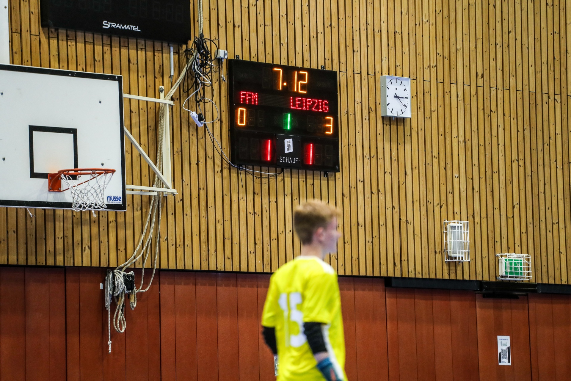 [{"clang_id":1,"value":"Basketballhalle. Scoreboard zeigt 7:12, FFM vs. Leipzig. Basketballkorb links, Spieler mit gelbem Trikot rechts im Hintergrund. Holzvertäfelung an der Wand."}]