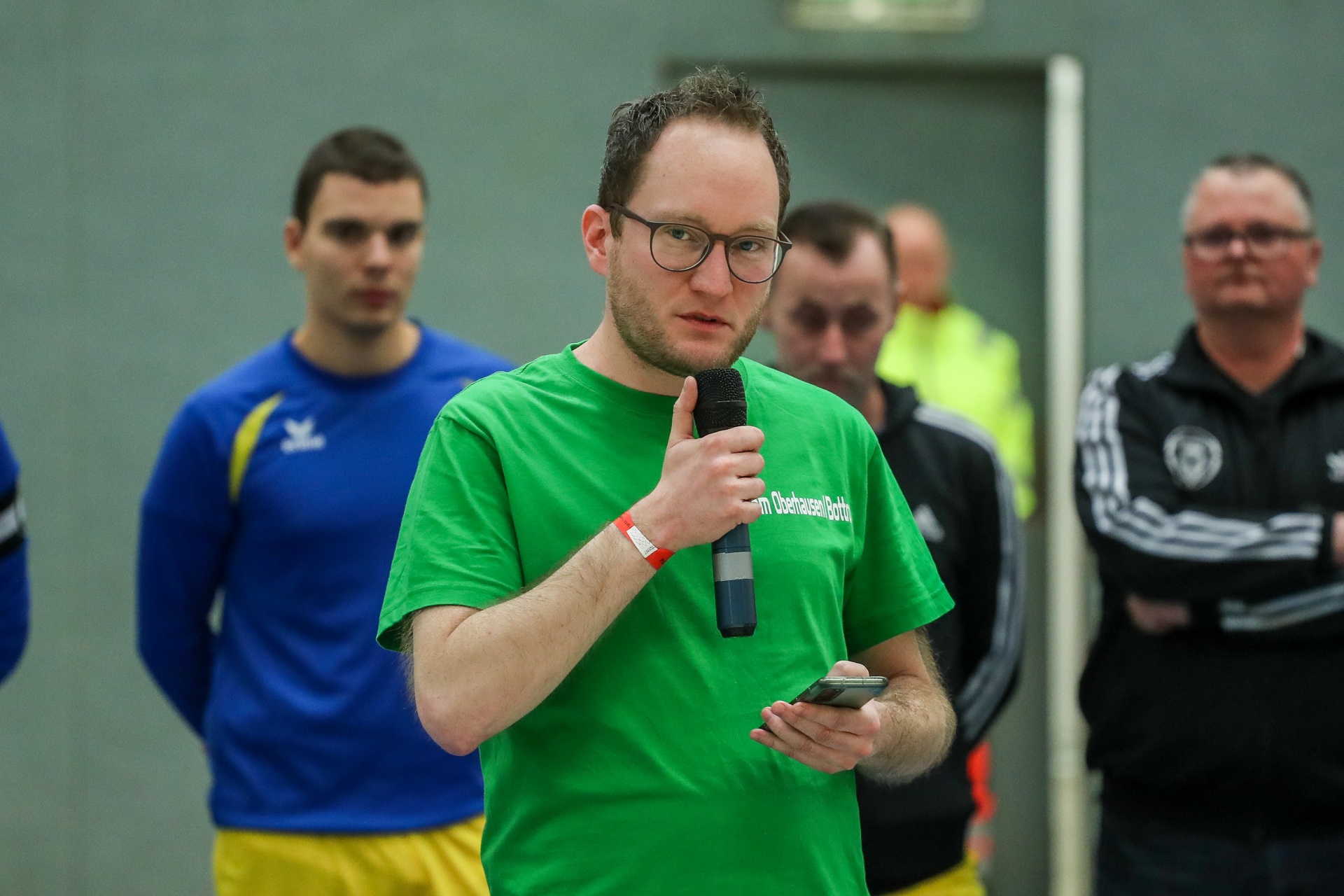 [{"clang_id":1,"value":"Mann mit Mikrofon spricht in Sporthalle. Im Hintergrund weitere Männer, einige in Sportkleidung, vor grauer Wand."}]