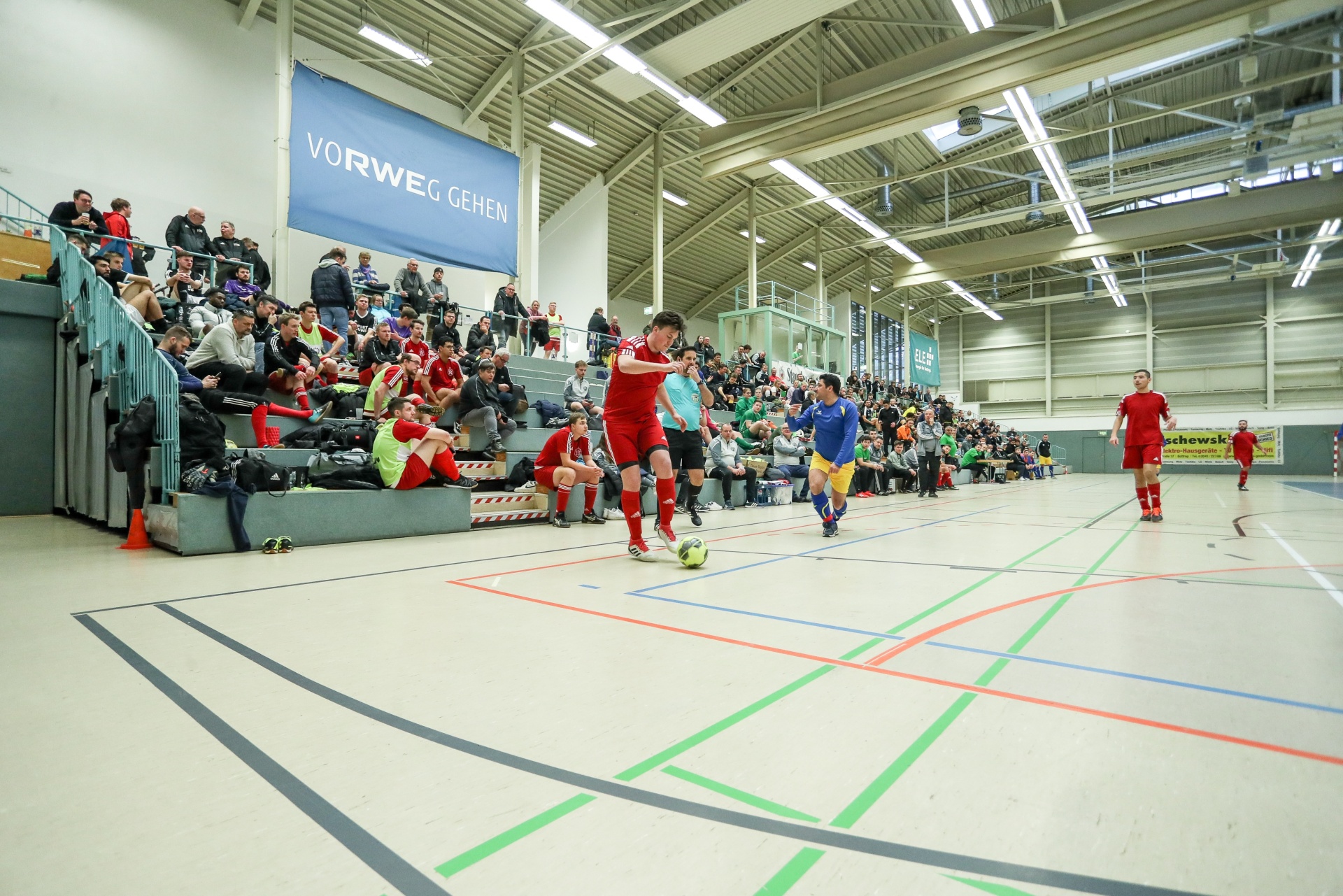 [{"clang_id":1,"value":"Indoor-Futsalspiel in einer Halle mit Zuschauertribüne. Spieler in roten und blauen Trikots kämpfen um den Ball. Grün und Grau markieren das Spielfeld."}]