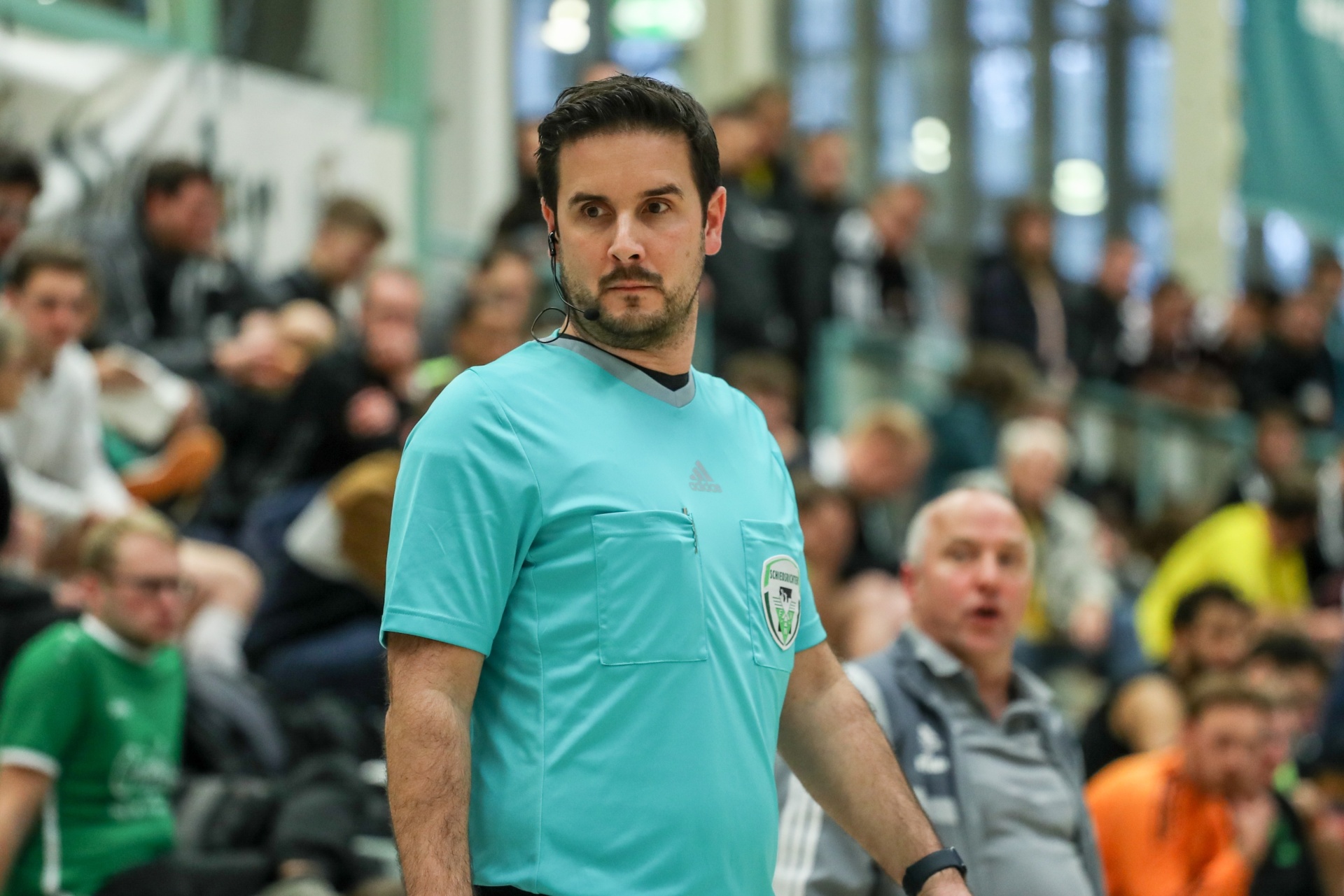 [{"clang_id":1,"value":"Handballschiedsrichter in türkisem Trikot mit Headset und Vereinsemblemen auf der Brust. Im Hintergrund verschwommene Zuschauer und Spieler."}]