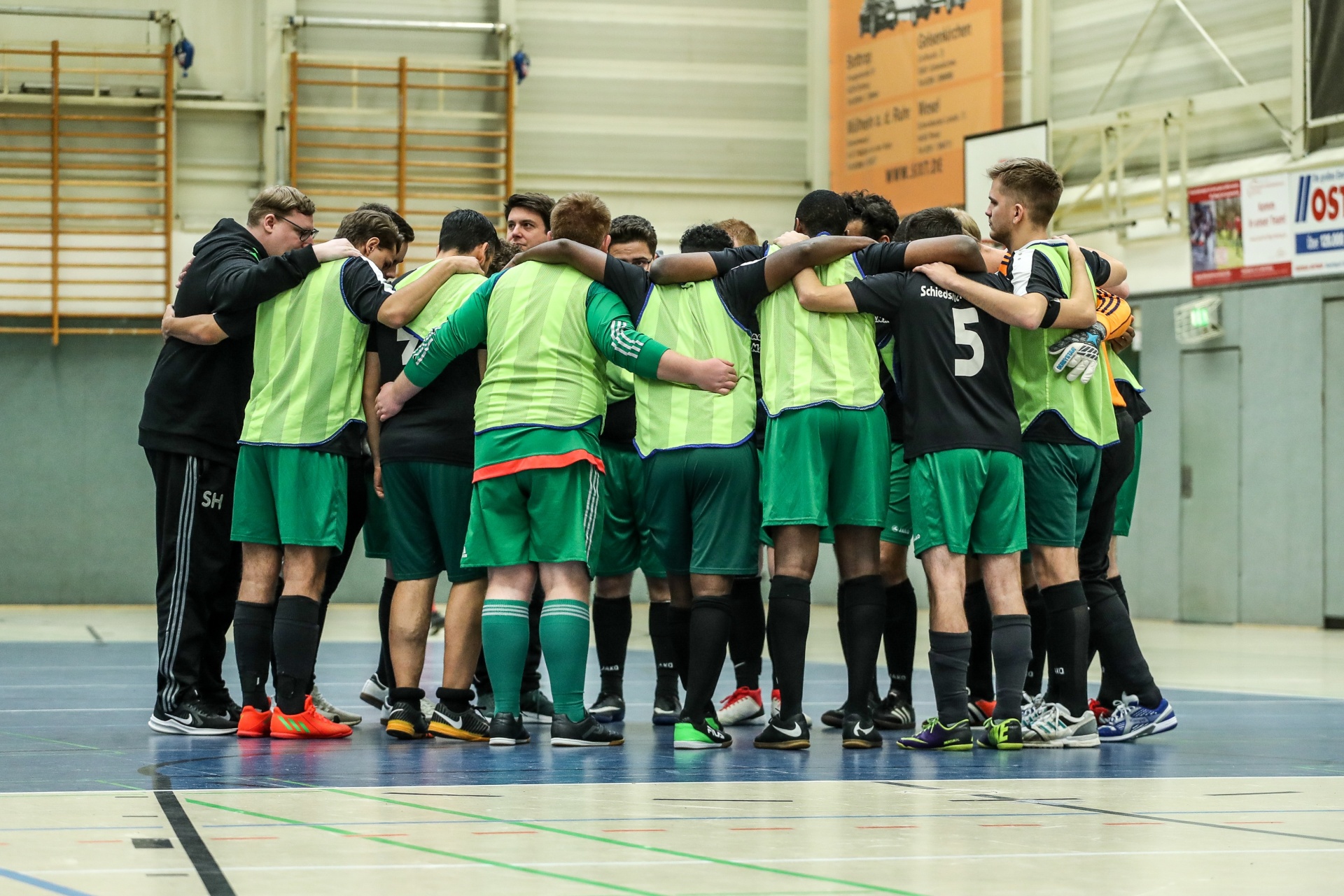 [{"clang_id":1,"value":"Handballteam im Kreis, überwiegend in grünen Trikots und Westen. Ein Trainer steht am Rand. Sporthalle im Hintergrund."}]