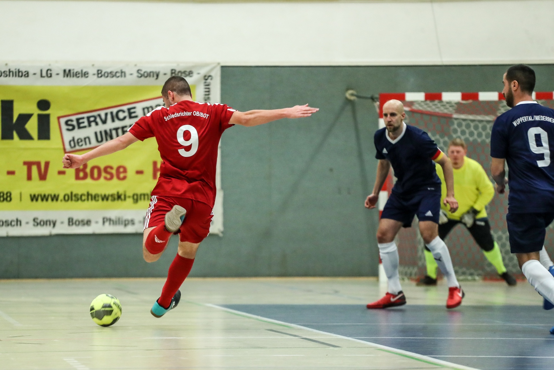 [{"clang_id":1,"value":"Futsalspiel: Zwei Spieler in roten Trikots mit Nummer 9 kämpfen um einen Ball auf dem Hallenfußballfeld. Im Hintergrund weitere Spieler und das Tor."}]