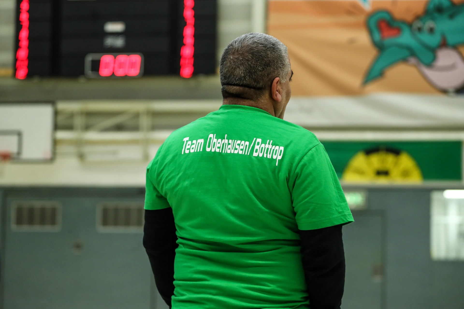 [{"clang_id":1,"value":"Mann mit grünem T-Shirt mit Aufschrift \"Team Oberhausen/Bottrop\" steht mit dem Rücken zum Betrachter in einer Sporthalle. Im Hintergrund eine Anzeigetafel."}]