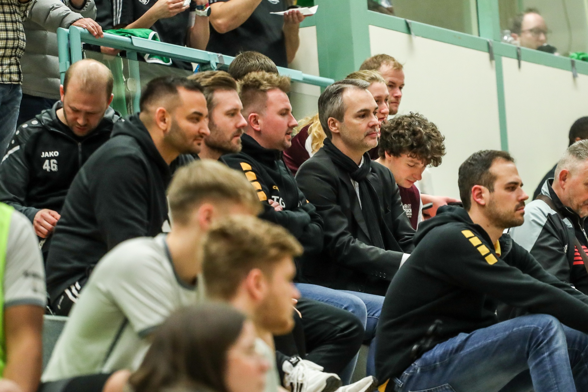 [{"clang_id":1,"value":"Handballteam sitzt auf einer Bank am Spielfeldrand. Männer unterschiedlichen Alters, einige mit Trainingsanzügen. Zuschauerränge im Hintergrund."}]