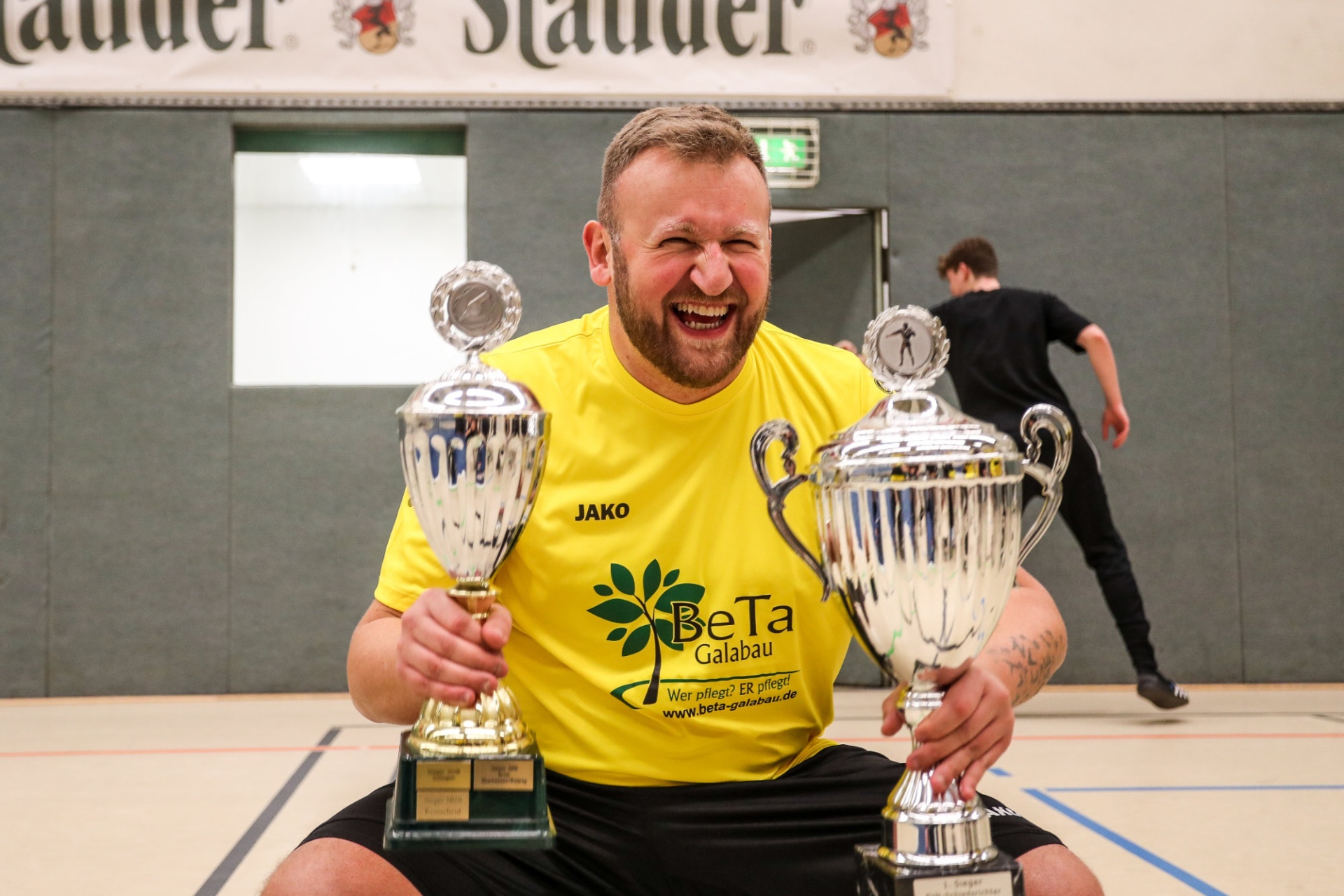 [{"clang_id":1,"value":"Mann mit Pokalen. Breit lächelnder Mann in gelbem T-Shirt mit Sponsorenlogo umgeben von sechs silbernen Pokalen in einer Sporthalle. Im Hintergrund verschwommen eine weitere Person."}]