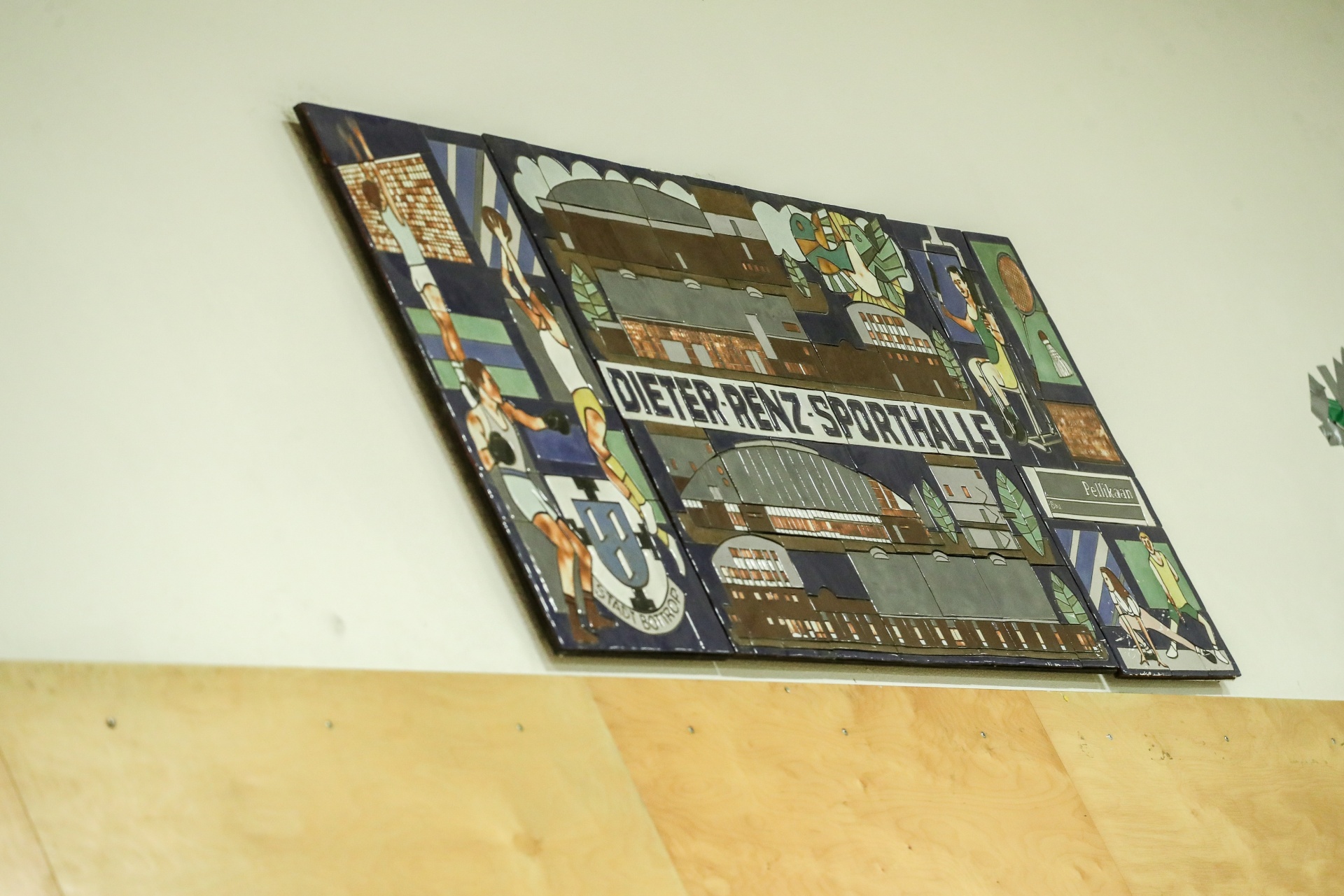 [{"clang_id":1,"value":"Wandbild mit stilisierten Darstellungen von Gebäuden, Sportlern und der Aufschrift \"DIETER-RENZ-SPORTHALLE\". Hängt an einer hellen Wand über einem Holzpaneel."}]