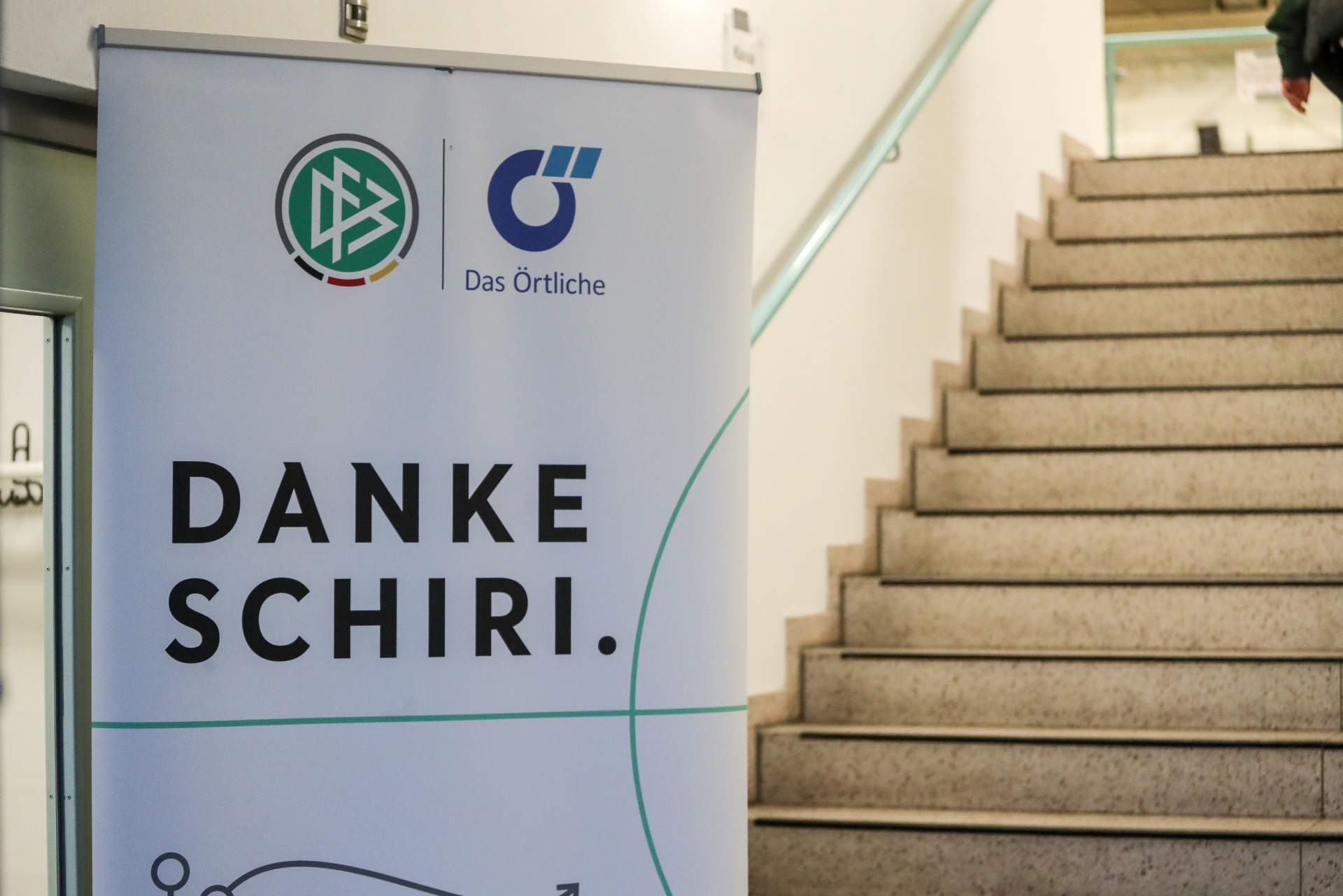 [{"clang_id":1,"value":"Werbebanner mit Aufschrift \"DANKE SCHIRI.\" und Logos von \"Das Örtliche\" vor einer hellen Treppe im Innenraum."}]