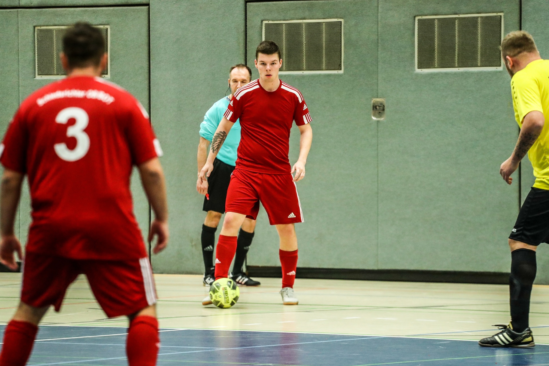 [{"clang_id":1,"value":"Fußballspieler in roten und gelben Trikots auf blauem Hallenboden. Ein Schiedsrichter steht im Hintergrund. Ein Fußball liegt auf dem Boden."}]