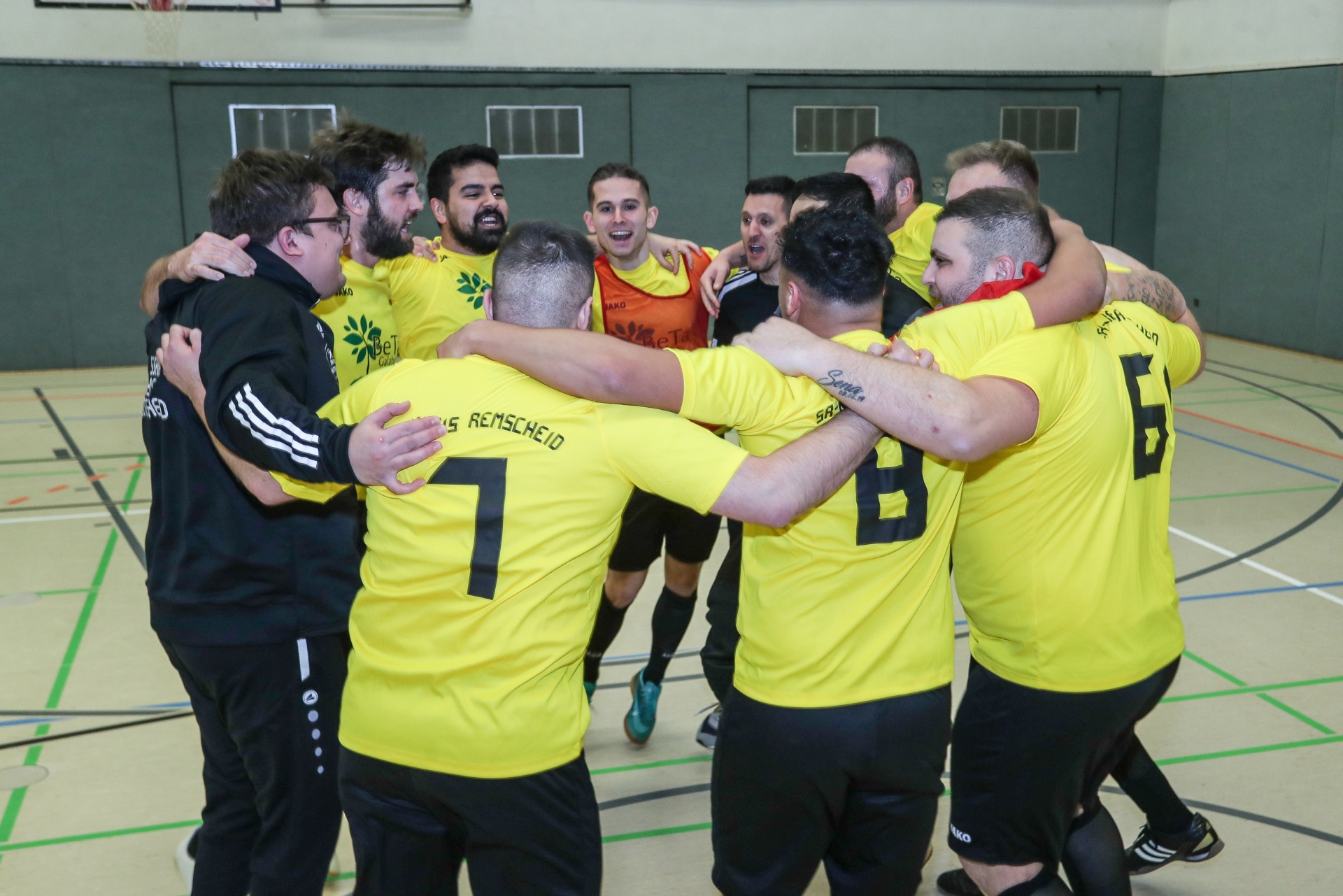 [{"clang_id":1,"value":"Handballspieler in gelben Trikots feiern im Kreis, umringt von Trainern und weiteren Teammitgliedern in einem Turnhalle."}]