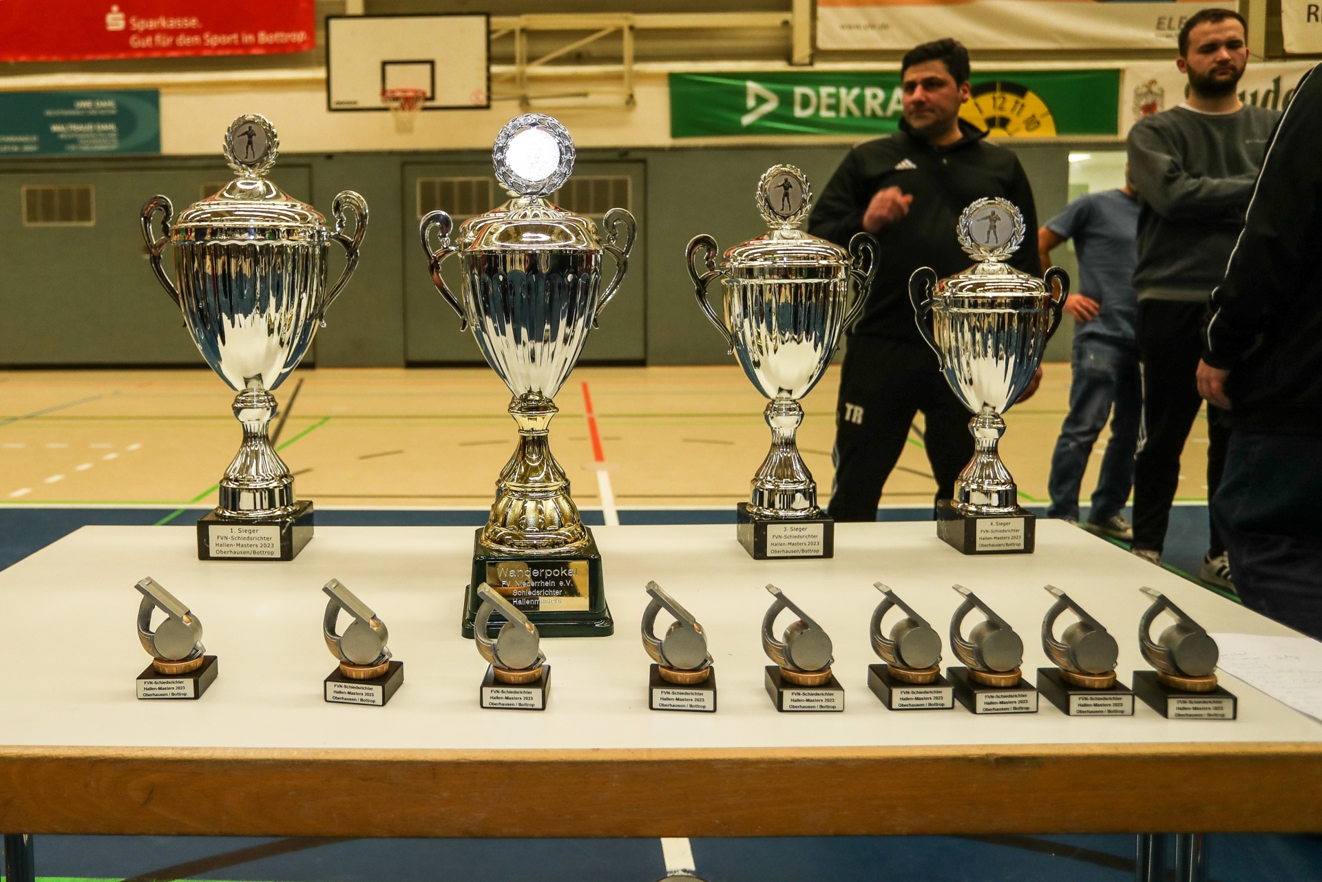 [{"clang_id":1,"value":"Pokale stehen auf einer mit weißem Tuch bedeckten Tisch in einer Sporthalle. Im Hintergrund Personen und Werbebanner."}]
