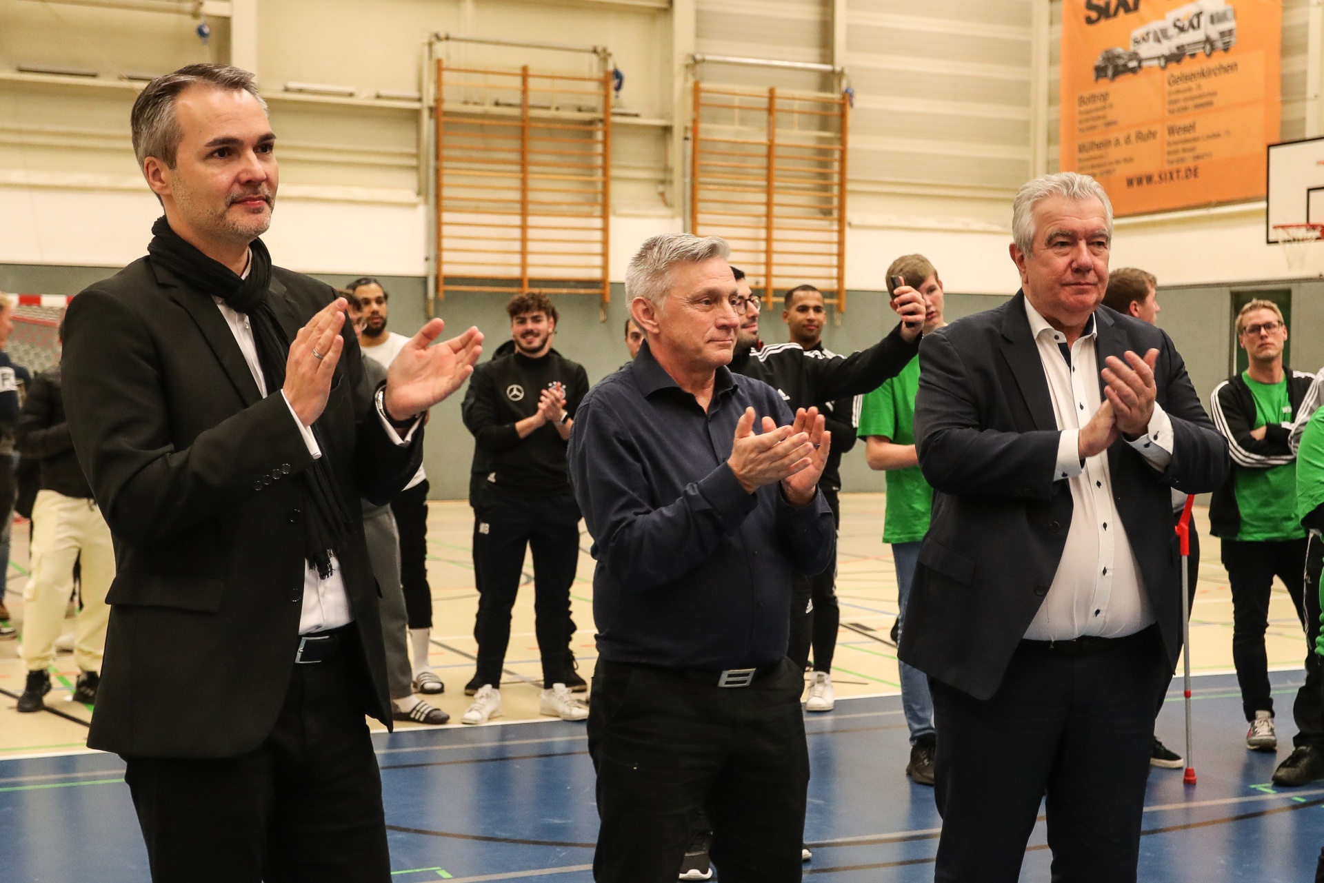 [{"clang_id":1,"value":"Drei Männer in Anzügen klatschen in einer Sporthalle. Im Hintergrund stehen junge Menschen. Wandbanner mit Text sind erkennbar."}]