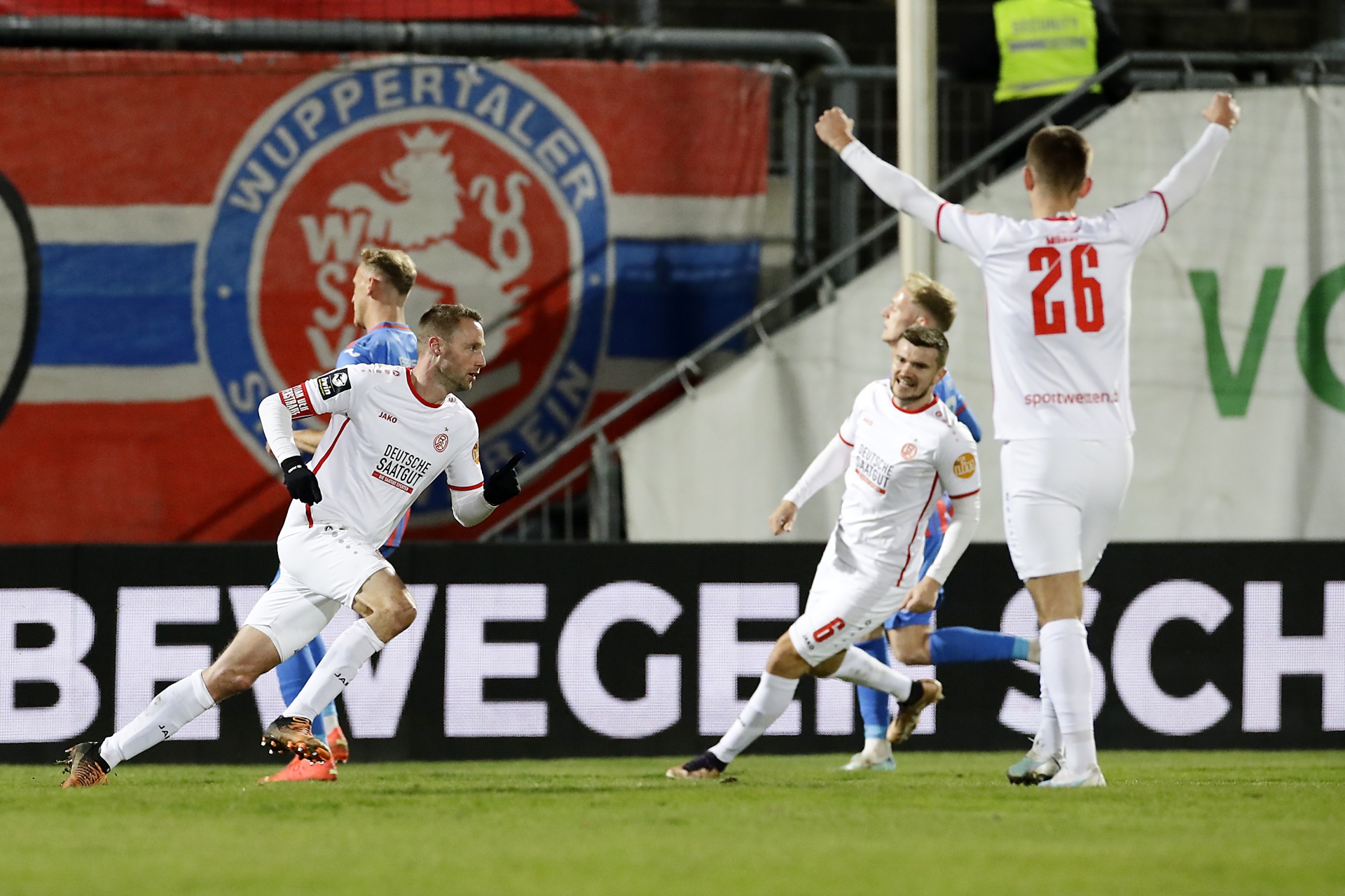 [{"clang_id":1,"value":"Fußballspieler feiern auf dem Rasen. Im Hintergrund ein Banner mit der Aufschrift \"WUPPERTALER\". Spieler tragen rot-weiße Trikots, ein Spieler die Nummer 26."}]