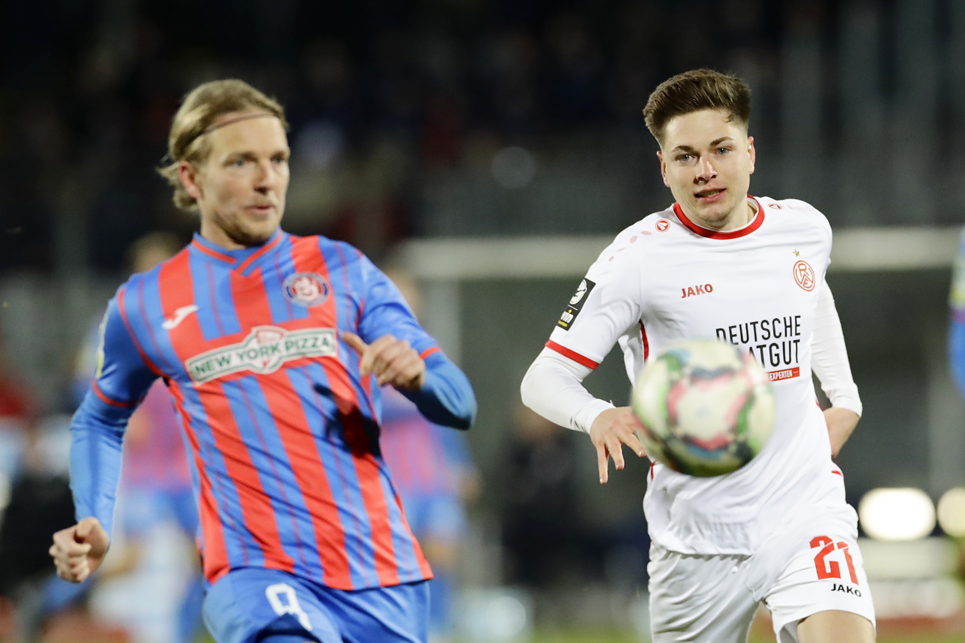 [{"clang_id":1,"value":"Zwei Fußballspieler auf dem Rasen. Rechts ein Spieler in weißen Trikots mit der Nummer 21. Links ein Spieler mit rot-blau gestreiftem Trikot und der Nummer 9."}]