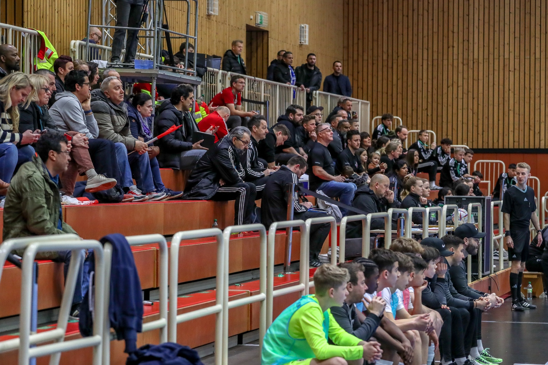 [{"clang_id":1,"value":"Zuschauerreihen in einer Sporthalle. Menschen sitzen und stehen auf orangefarbenen Bänken, teilweise vor weißem Geländer. Im Hintergrund weitere Zuschauer und eine Wand mit Holzvertäfelung."}]
