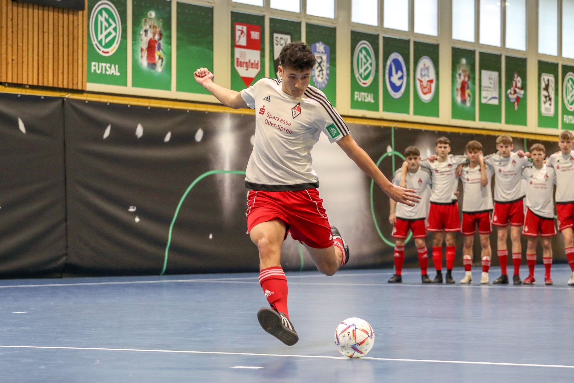 [{"clang_id":1,"value":"Futsalspieler in roter und weißer Ausrüstung schießt Ball in Sporthalle. Im Hintergrund weitere Spieler und Werbebanner."}]