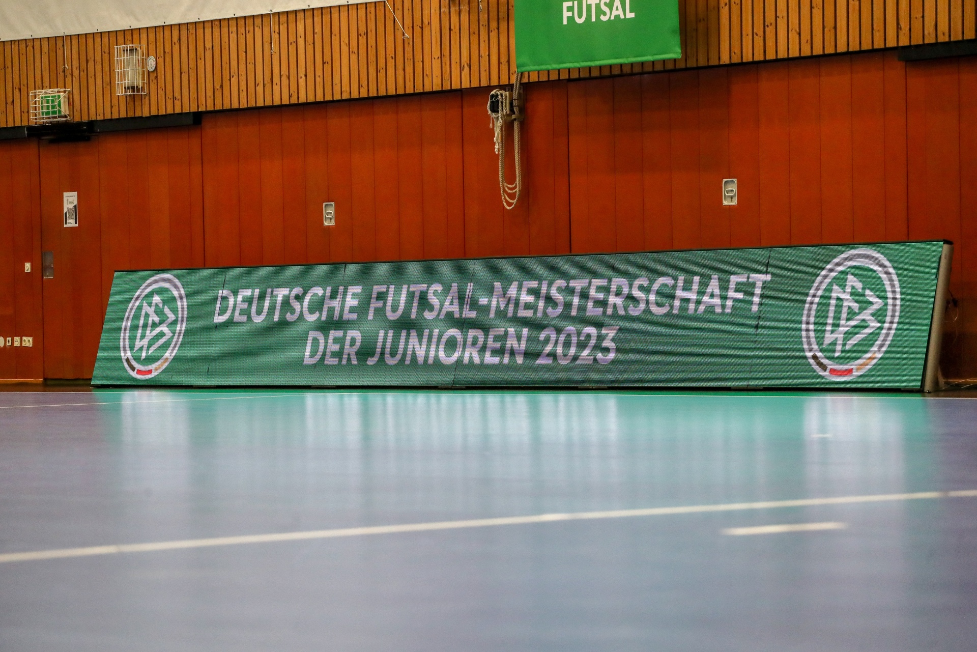 [{"clang_id":1,"value":"Futsal-Halle mit Werbebanner: \"Deutsche Futsal-Meisterschaft der Junioren 2023\". Grauer Hallenboden, braune Wandpaneele, grünes \"Futsal\"-Schild."}]