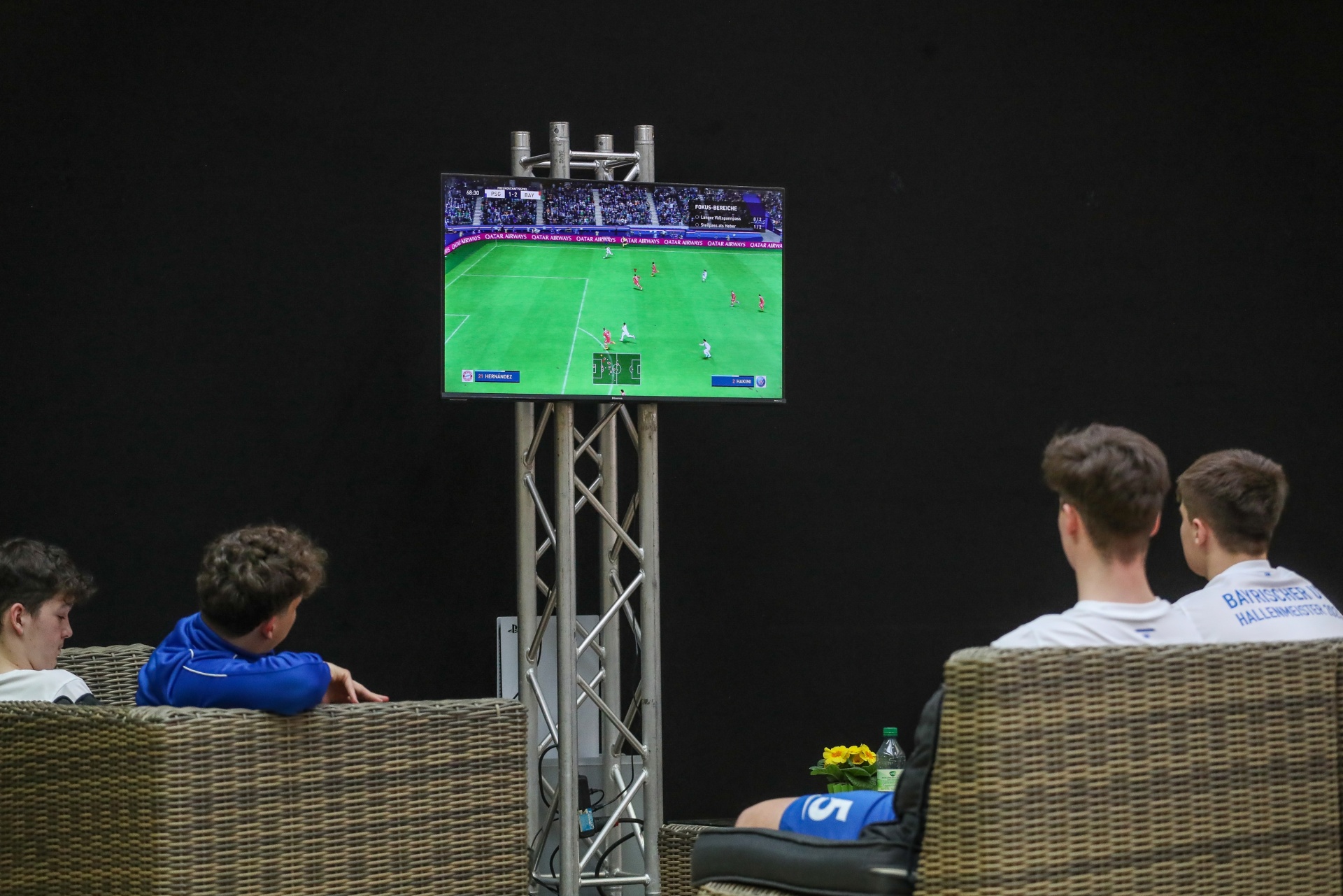 [{"clang_id":1,"value":"Fußballspiel auf großem Bildschirm. Vier junge Männer sitzen auf Rattanmöbeln und schauen zu. Getränke und Blumen stehen in der Nähe."}]