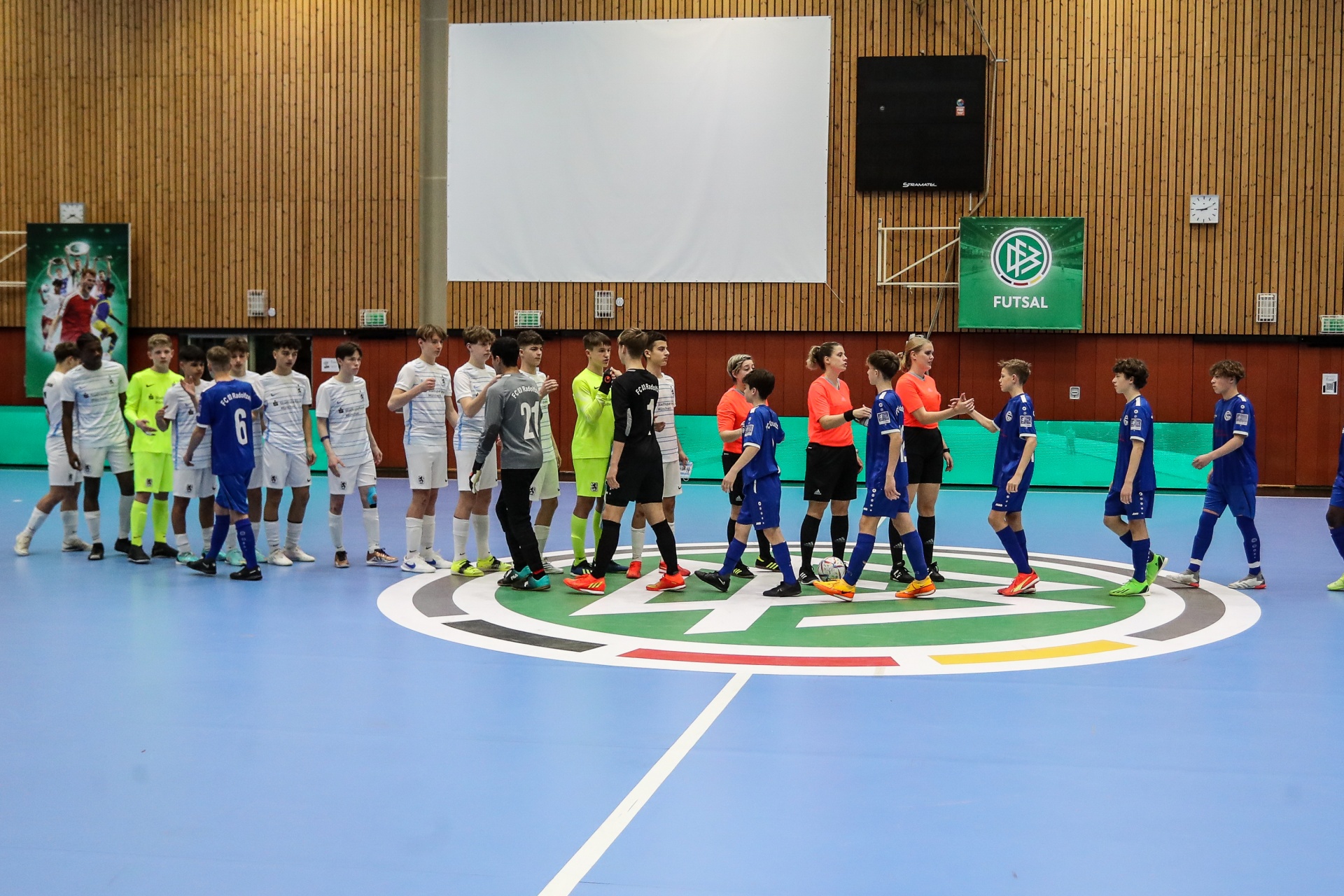 [{"clang_id":1,"value":"Futsal-Halle: Zwei Mannschaften in farbigen Trikots stehen sich gegenüber, umgeben von Schiedsrichtern. Im Hintergrund eine hölzerne Wand und eine Leinwand."}]