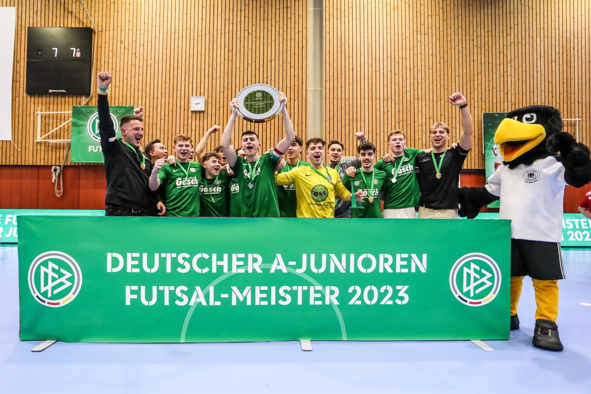 [{"clang_id":1,"value":"Futsal-Mannschaft mit Pokal. Zehn Spieler in grünen Trikots stehen vor einem grünen Banner mit der Aufschrift \"Deutscher A-Junioren Futsal-Meister 2023\". Rechts eine Maskottchenfigur."}]