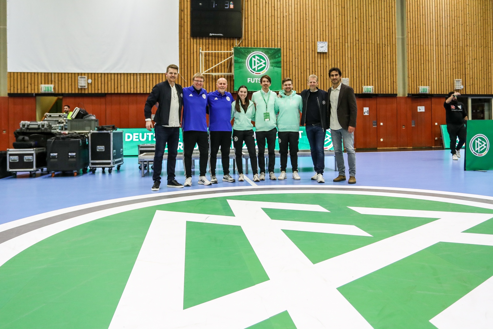 [{"clang_id":1,"value":"Gruppe von acht Personen steht auf einem Indoor-Sportboden mit grün-weißem Muster vor einer hölzernen Wand. Im Hintergrund befinden sich Geräte und eine Anzeigetafel."}]
