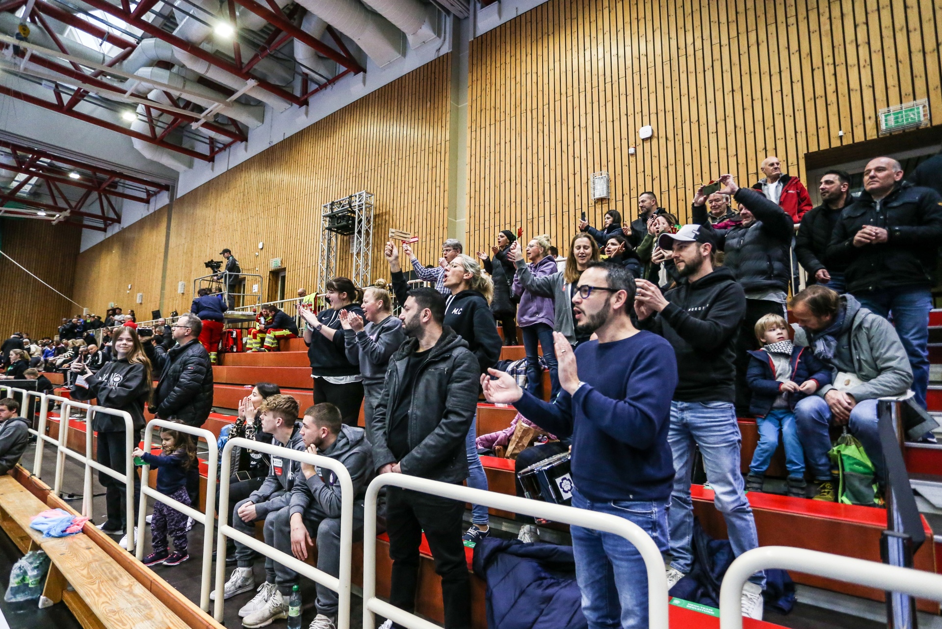 [{"clang_id":1,"value":"Zuschauer auf einer Tribüne in einer Sporthalle. Viele Menschen sitzen oder stehen, beobachten ein Ereignis im Fokusbereich. Holzvertäfelung im Hintergrund."}]