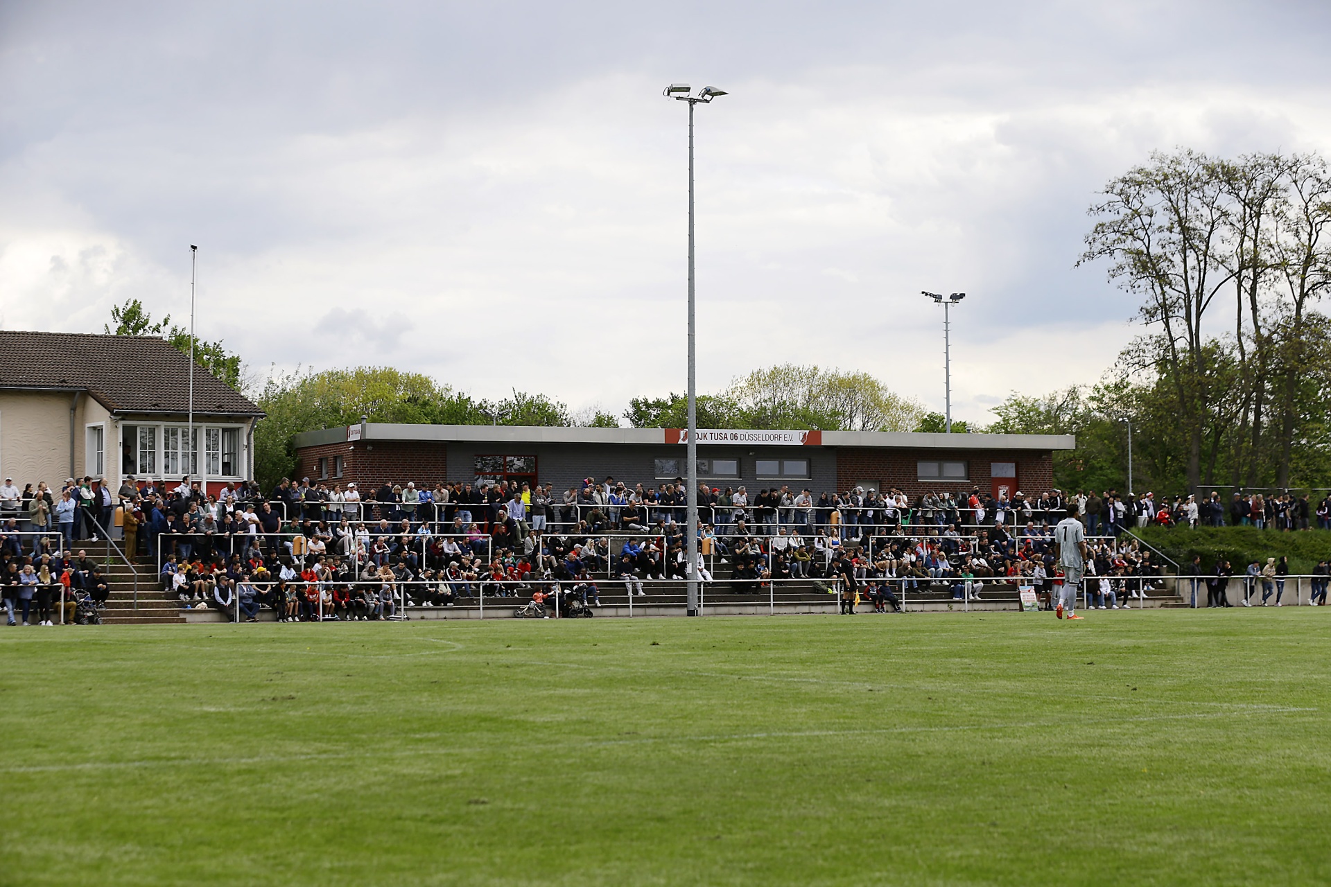 [{"clang_id":1,"value":"Fußballstadion mit Zuschauerrängen. Rasenplatz im Vordergrund, Gebäude mit Zuschauern im Hintergrund, Lichtmasten. Bewölkter Himmel."}]