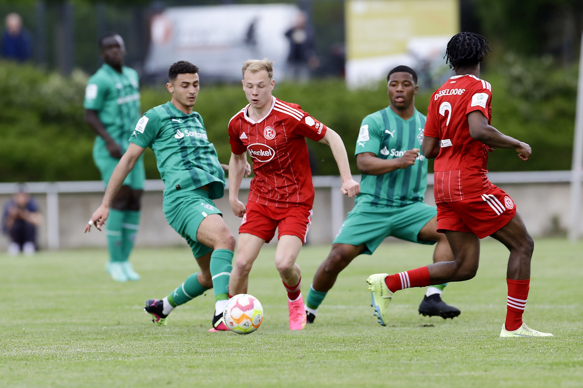 [{"clang_id":1,"value":"Fußballspiel: Drei Spieler in rot und drei in grün kämpfen um den Ball auf einem grünen Rasenplatz. Im Hintergrund Zuschauer und Bäume."}]