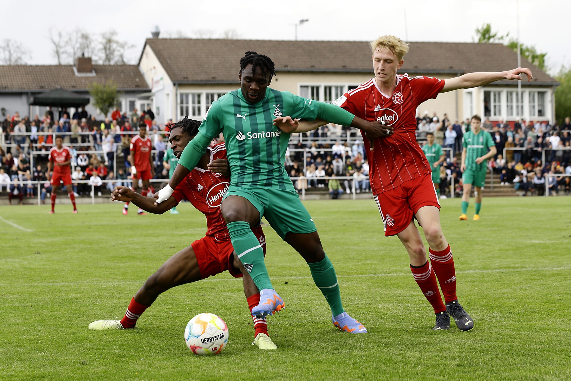 [{"clang_id":1,"value":"Fußballspiel: Drei Spieler kämpfen um den Ball auf grünem Rasen. Ein Spieler in Grün und zwei in Rot. Zuschauer im Hintergrund."}]