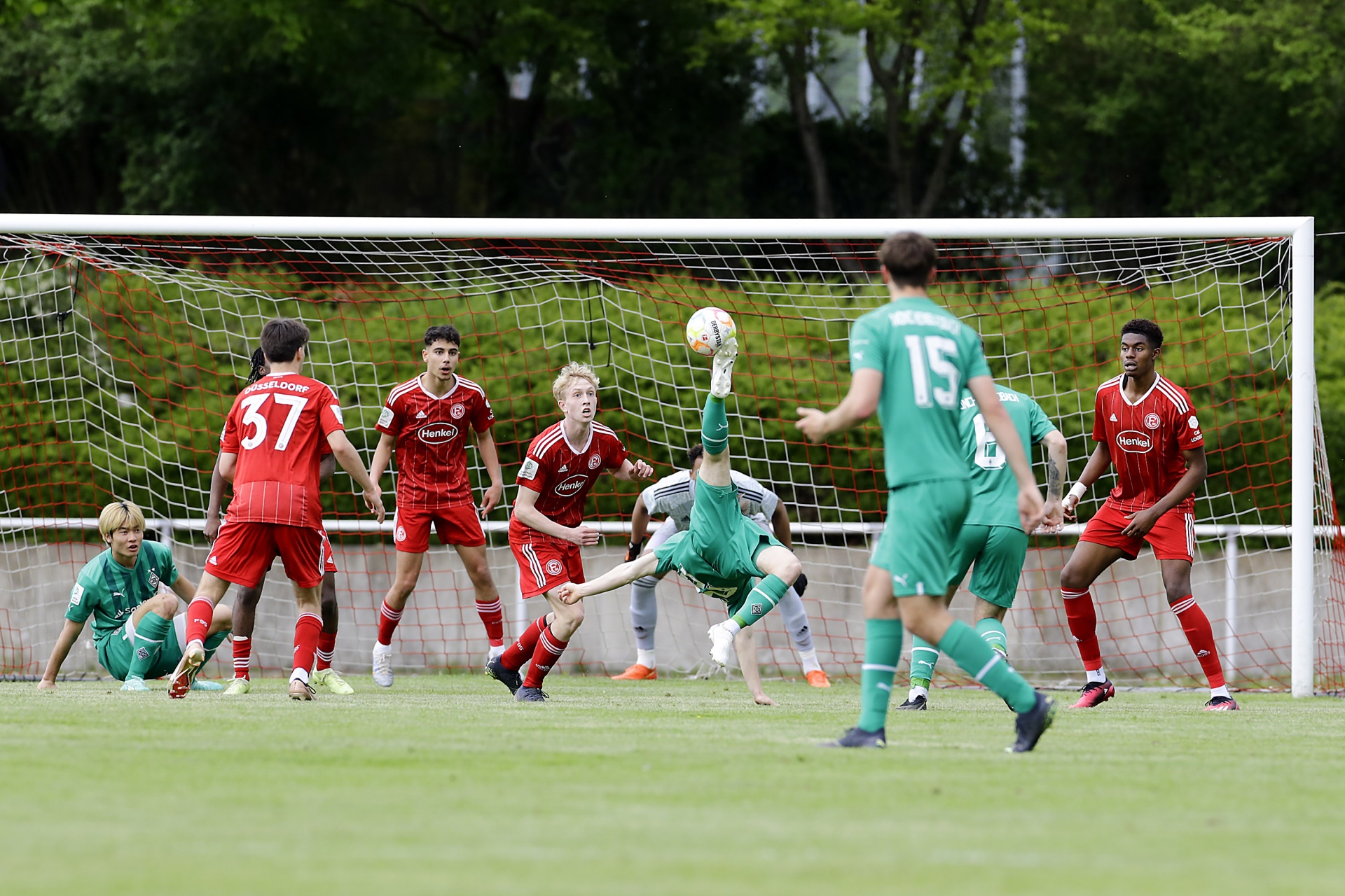 [{"clang_id":1,"value":"Fußballspiel: Spieler in roten und türkisen Trikots kämpfen um den Ball vor dem Tor. Im Hintergrund Zuschauer und Bäume."}]
