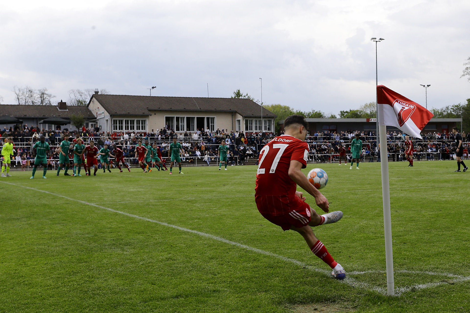 [{"clang_id":1,"value":"Fußballspieler in rotem Trikot schießt Ecke. Im Hintergrund Zuschauer und Gebäude auf grünem Rasen."}]