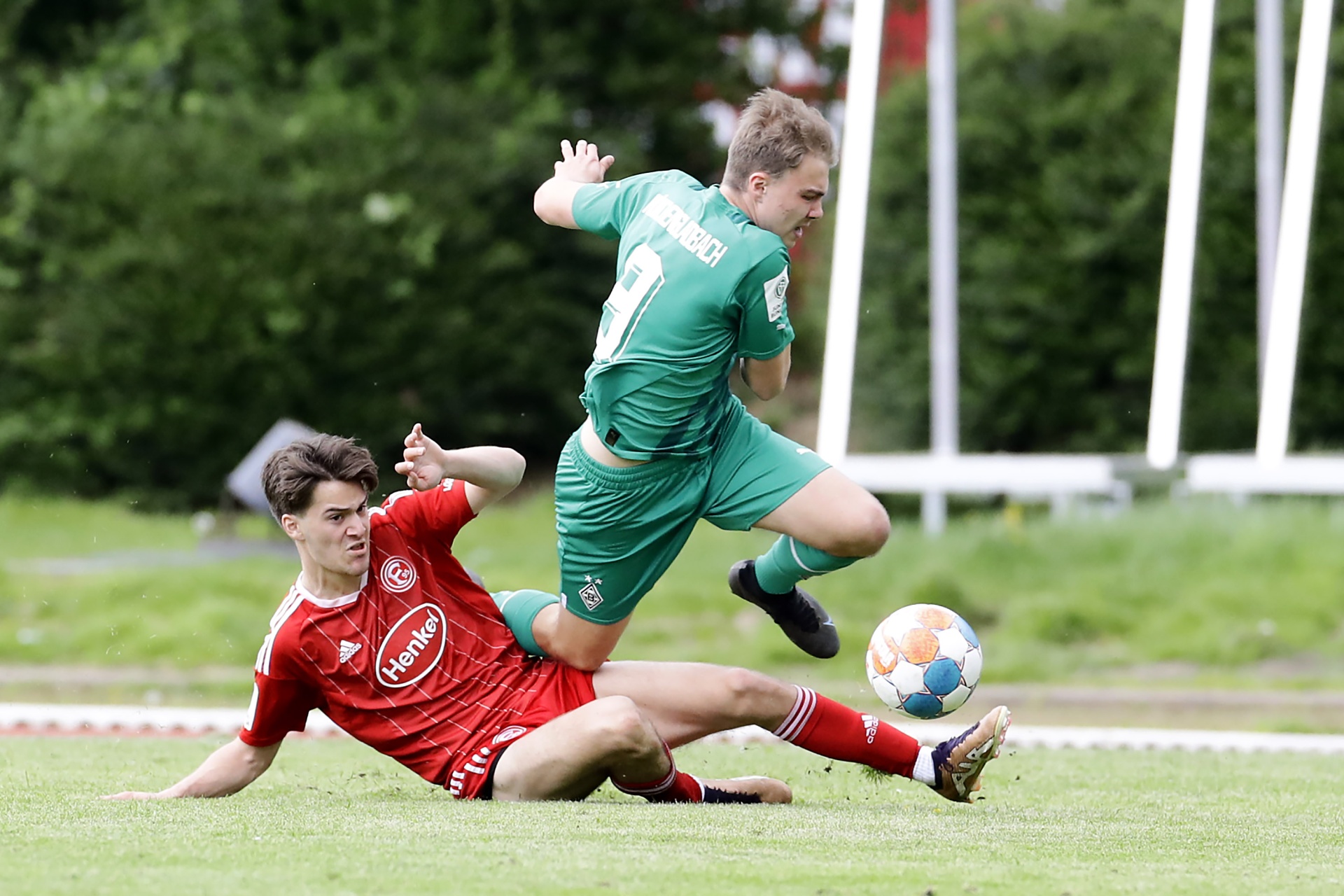 [{"clang_id":1,"value":"Zwei Fußballspieler im Spiel. Ein Spieler in Rot rutscht, der andere in Grün springt über ihn und den Ball. Hintergrund: Grünfläche und Zaun."}]