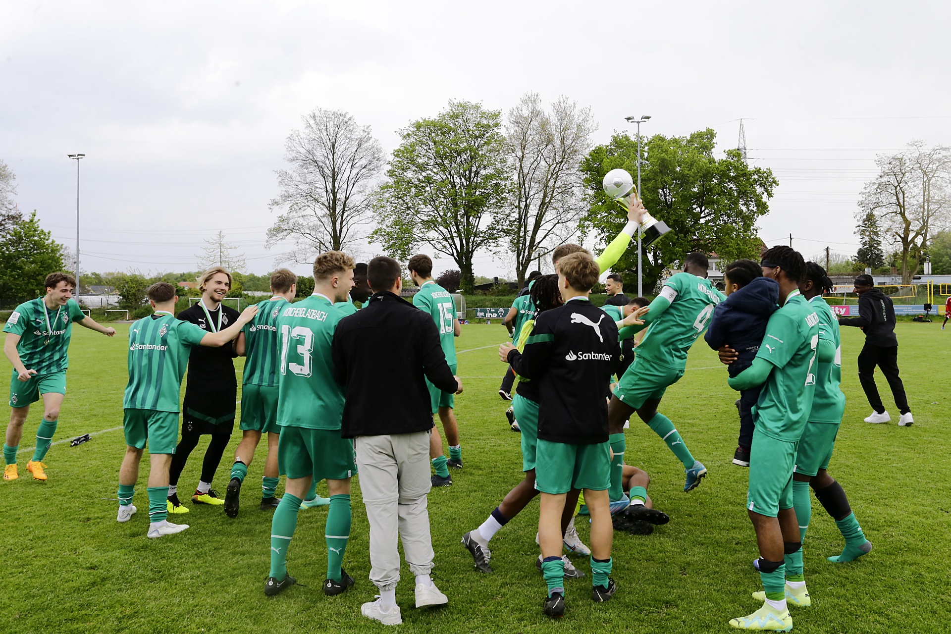 [{"clang_id":1,"value":"Fußballmannschaft in grünen Trikots feiert auf dem Spielfeld. Einige Spieler springen und jubeln, andere stehen im Kreis. Im Hintergrund Bäume und ein Fußballfeld."}]