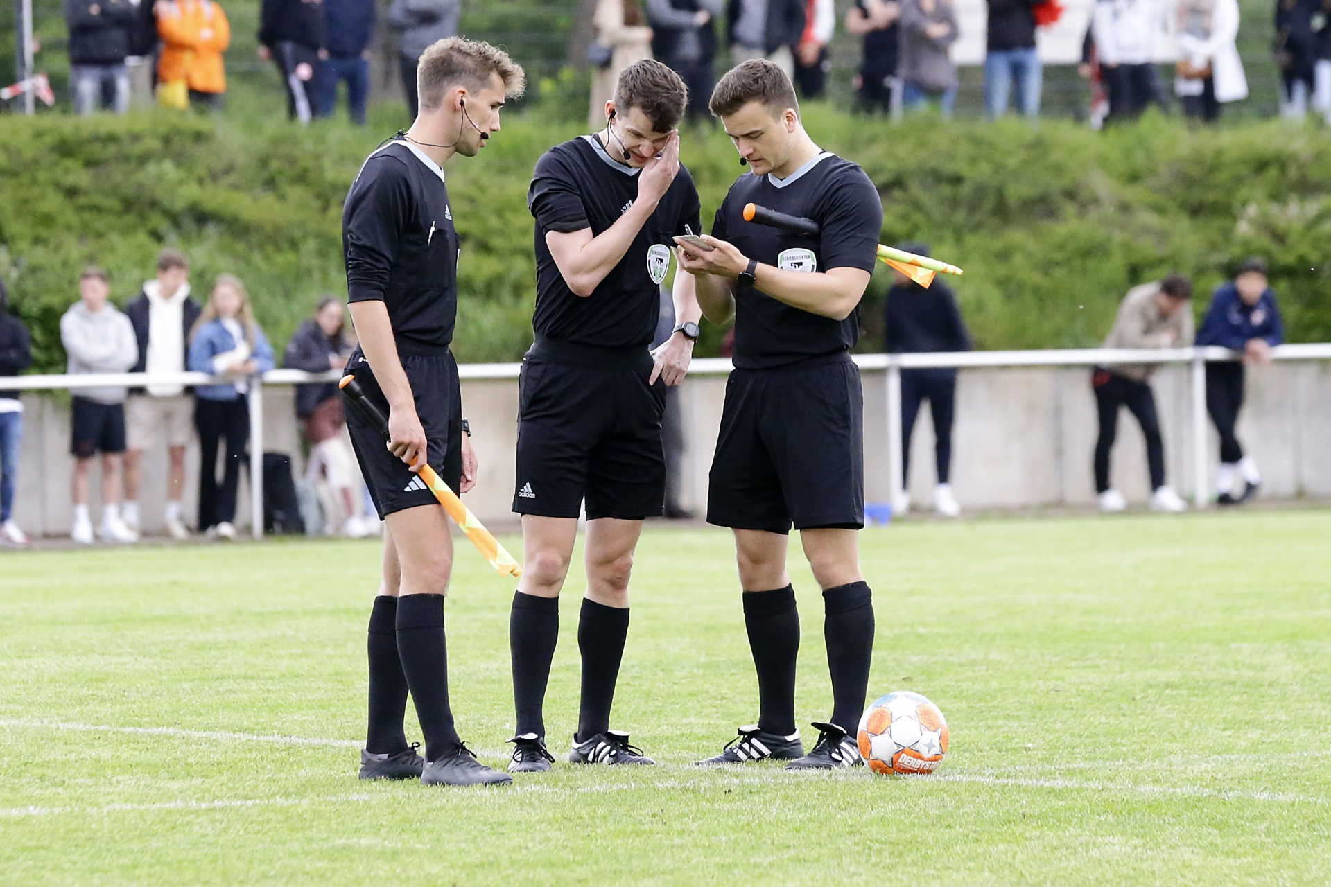 [{"clang_id":1,"value":"Drei Fußballschiedsrichter stehen auf einem grünen Rasen. Der mittlere hält ein Tablet, die anderen jeweils eine Flagge. Im Hintergrund Zuschauer und eine Absperrung."}]