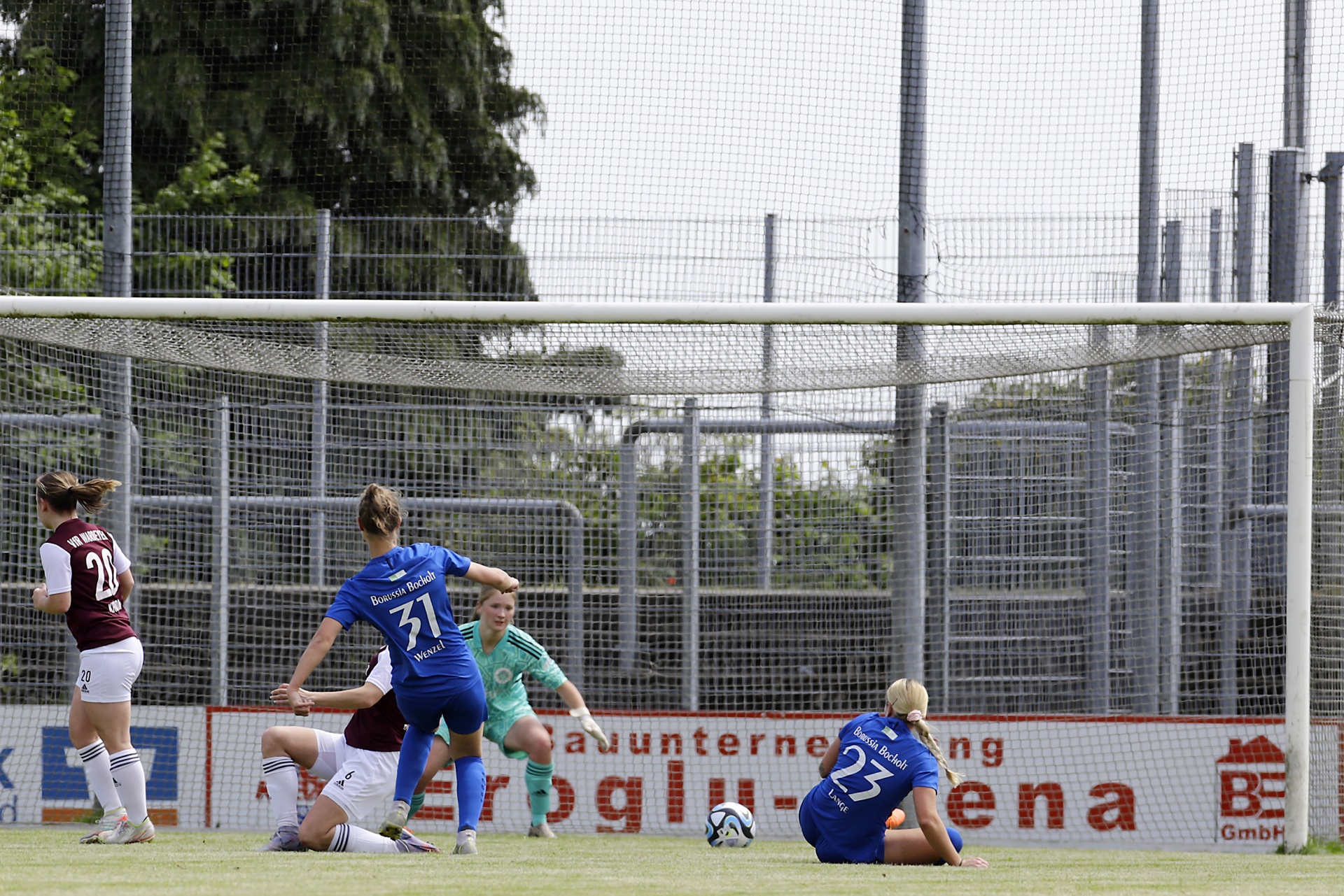 [{"clang_id":1,"value":"Fußballspiel: Spielerinnen in roten und blauen Trikots kämpfen um den Ball vor einem Metalltor und einer Zuschauertribüne. Himmel im Hintergrund."}]