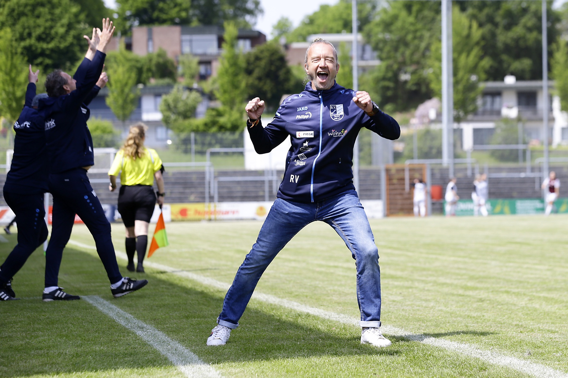 [{"clang_id":1,"value":"Fußballfeld. Mann in Trainingsjacke und Jeans jubelt mit erhobenen Fäusten. Im Hintergrund weitere Personen und Zuschauerränge."}]