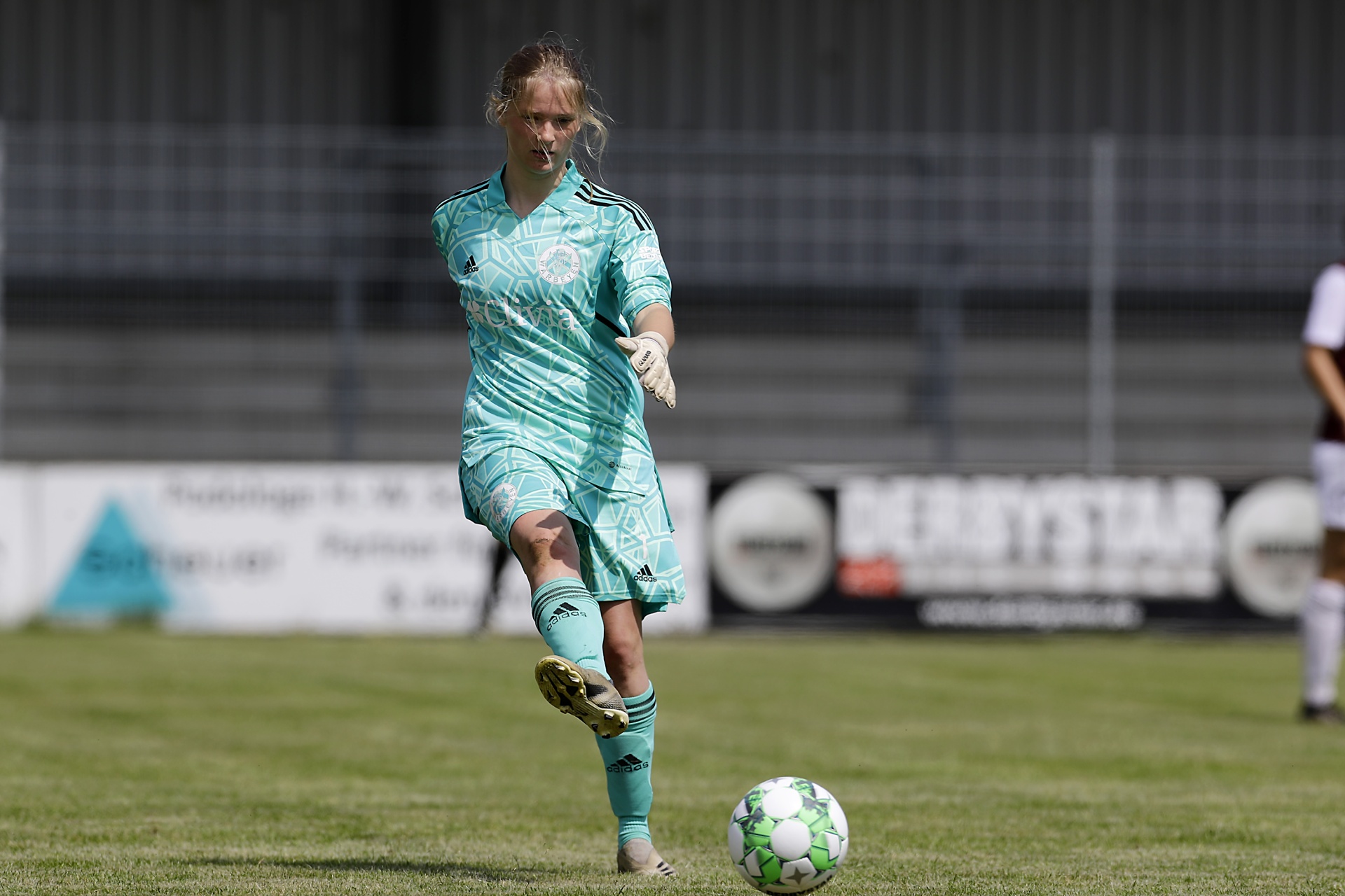 [{"clang_id":1,"value":"Fußballspielerin in türkisem Trainingsanzug schießt Ball auf grünem Rasen. Im Hintergrund Zaun und Werbebanner."}]
