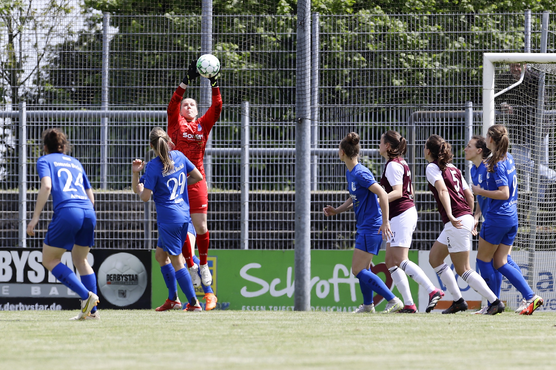 [{"clang_id":1,"value":"Fußballspiel: Torwartin in rot fängt Ball, umringt von Spielerinnen in blau und bordeauxrot vor Stadionbanden."}]