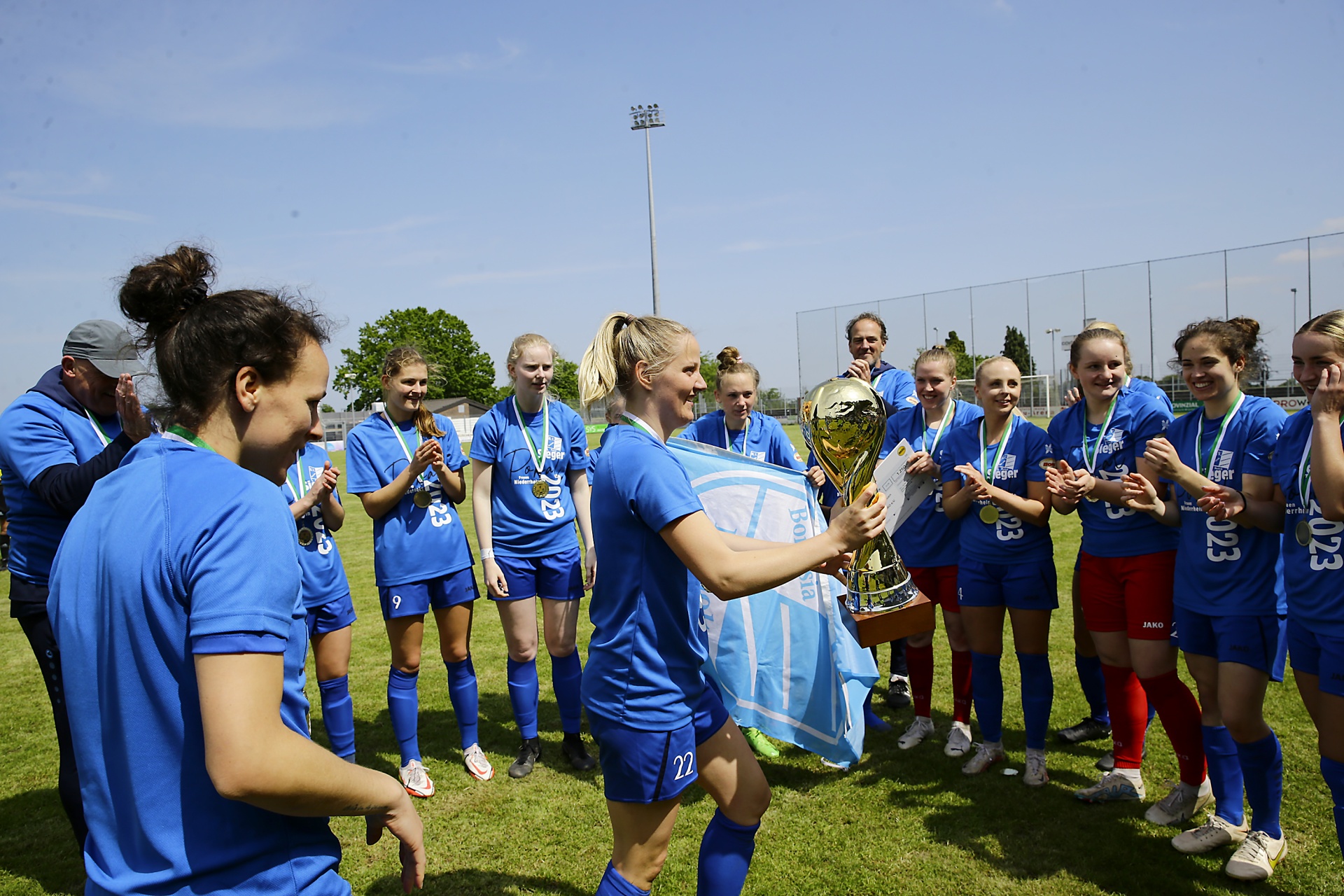 [{"clang_id":1,"value":"Fußballteam in blauen Trikots feiert Sieg mit Pokal auf dem Rasen. Spielerinnen umringen und berühren die Trophäe. Helle Umgebung, sonniger Tag."}]