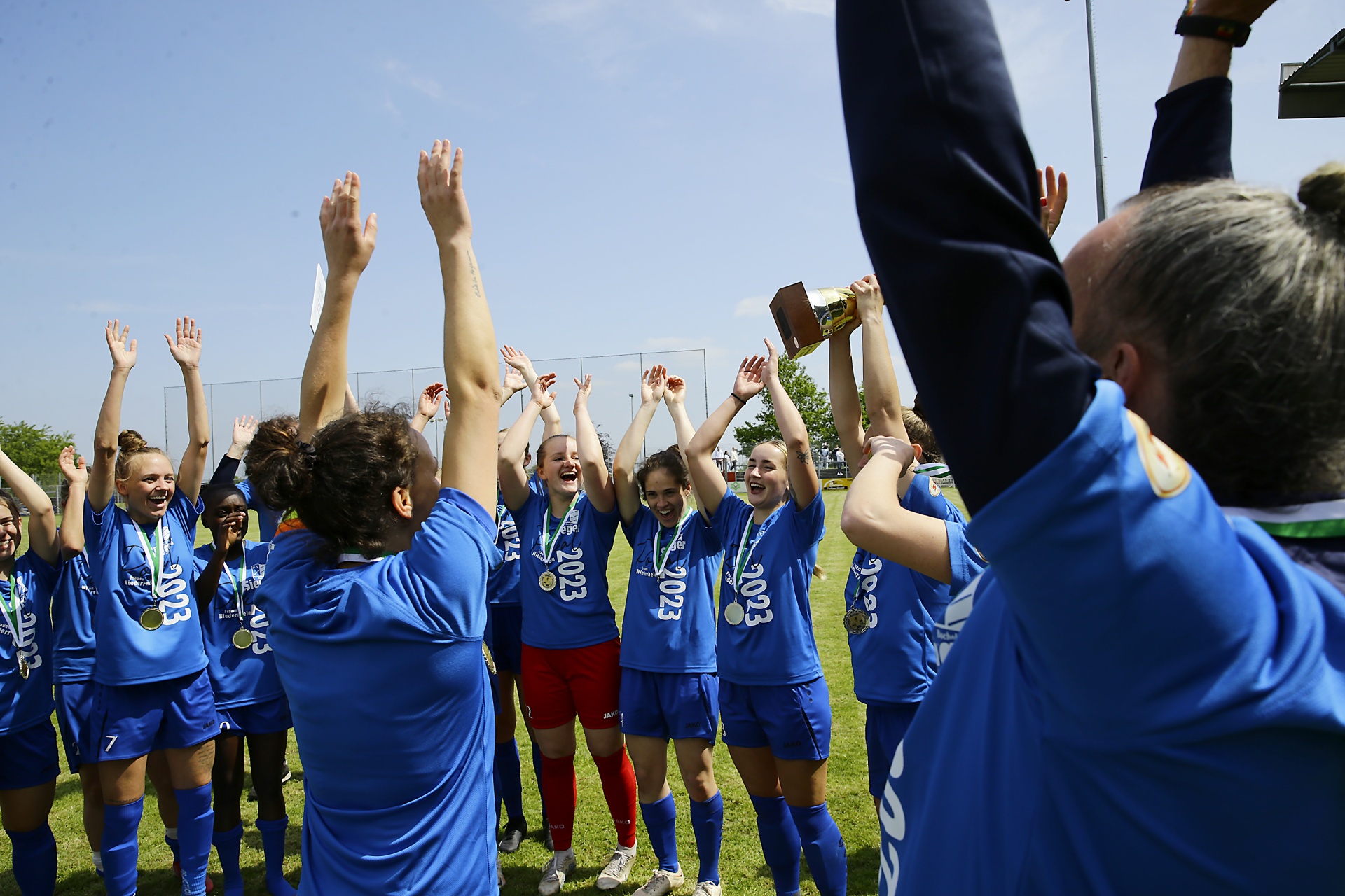 [{"clang_id":1,"value":"Fußballteam in blauen Trikots feiert Sieg mit Pokal und Medaillen. Spielerinnen halten Arme hoch, Trainer stehen im Hintergrund. Grünfläche, blauer Himmel."}]