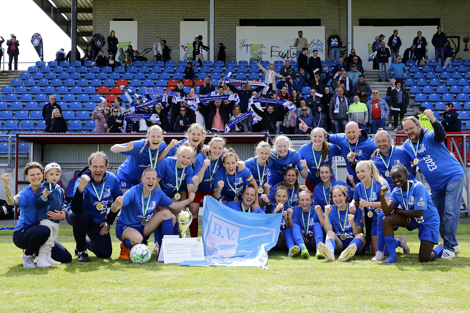 [{"clang_id":1,"value":"Frauenfußballteam in blauen Trikots feiert Sieg mit Pokal und Medaillen auf dem Rasen vor gefüllter Tribüne. Im Hintergrund Gebäude mit Zuschauern."}]