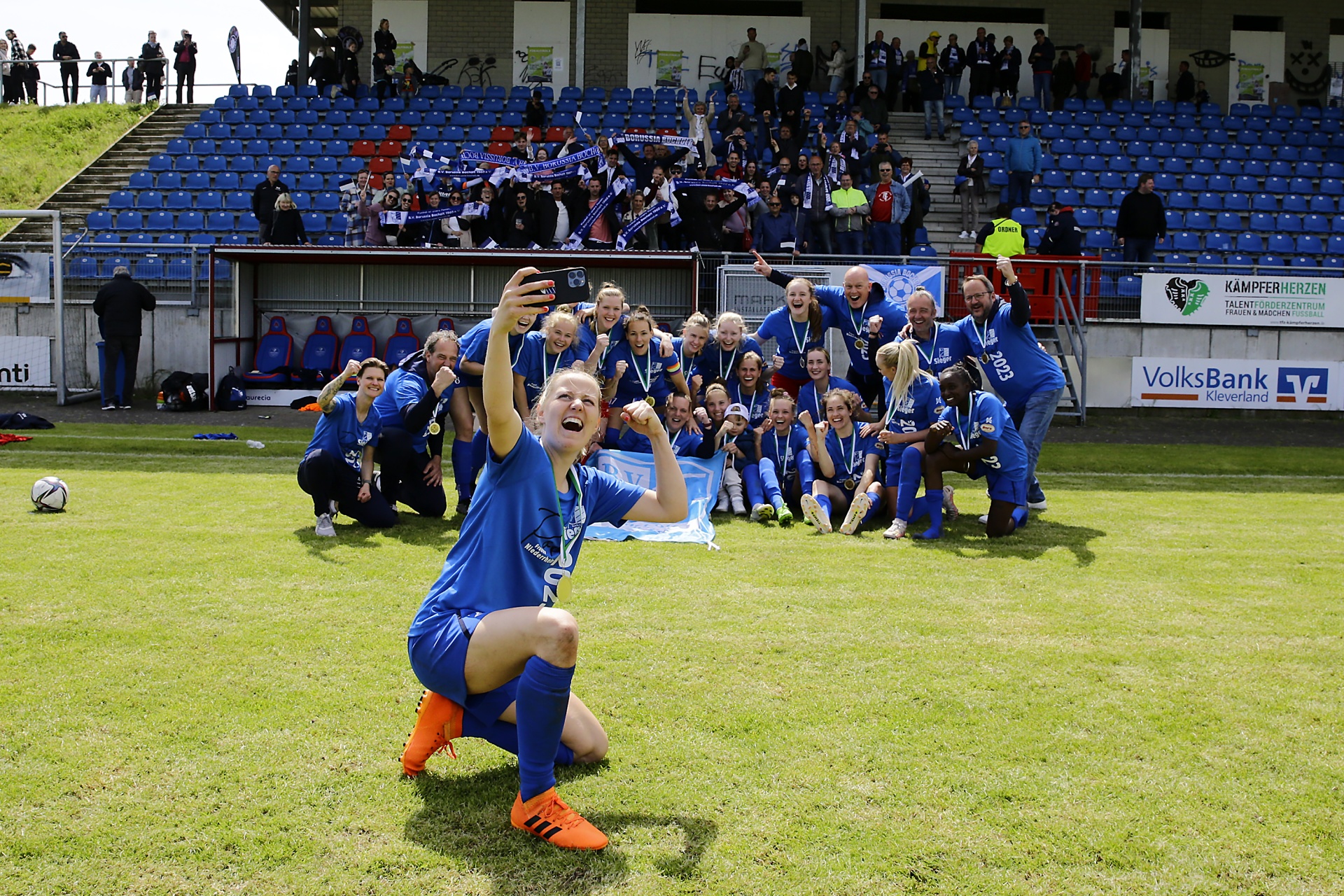 [{"clang_id":1,"value":"Frauenfußballteam in blauen Trikots jubelt auf dem Rasen vor gefüllter Tribüne, eine Spielerin kniet und hält Smartphone hoch. Siegerehrung."}]