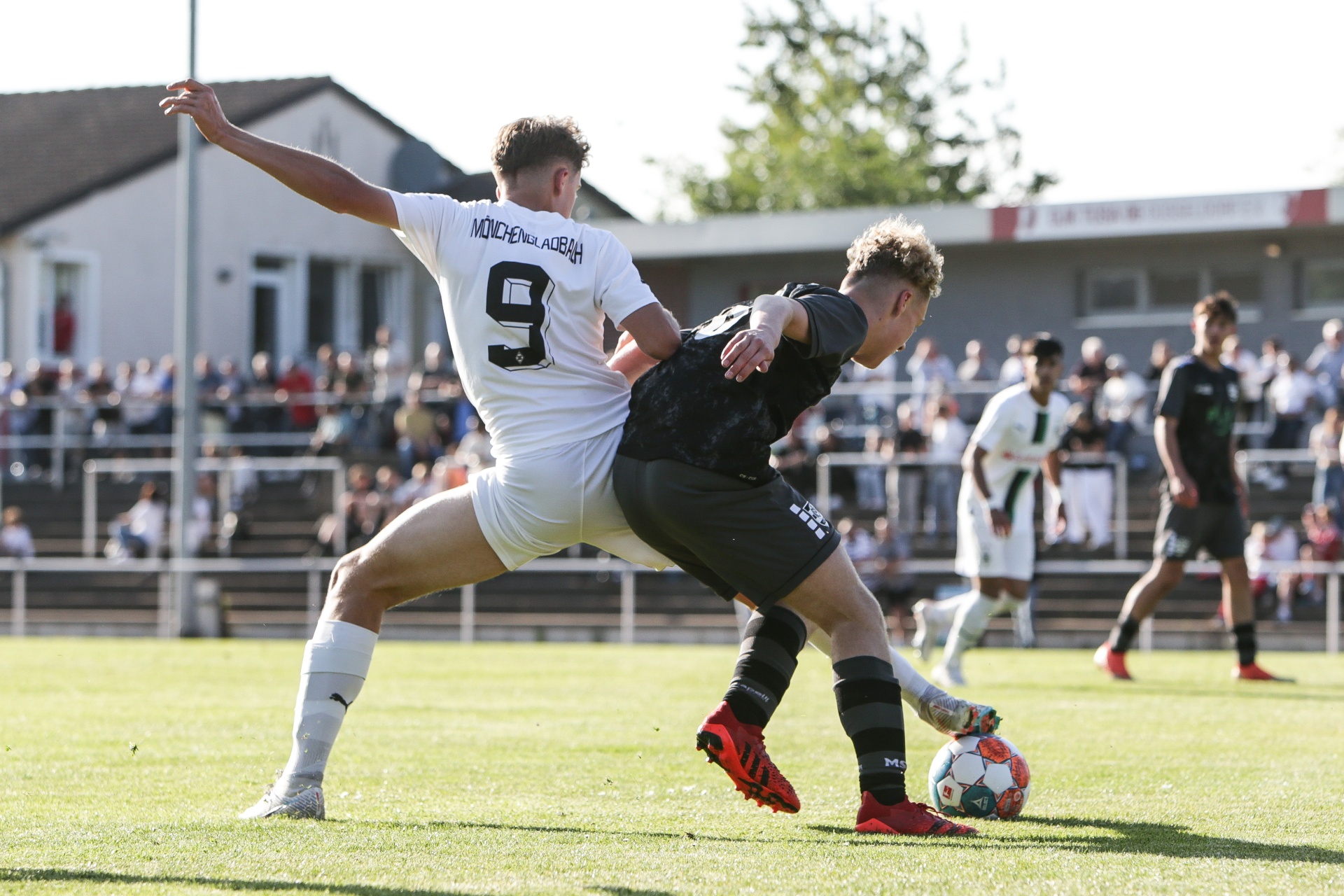 [{"clang_id":1,"value":"Zwei Fußballspieler kämpfen um den Ball. Ein Spieler in weißen Shorts und Trikot mit Nummer 9 wird von einem Spieler in schwarzen Shorts und rotem Schuhwerk angegriffen. Im Hintergrund Zuschauer und Gebäude."}]