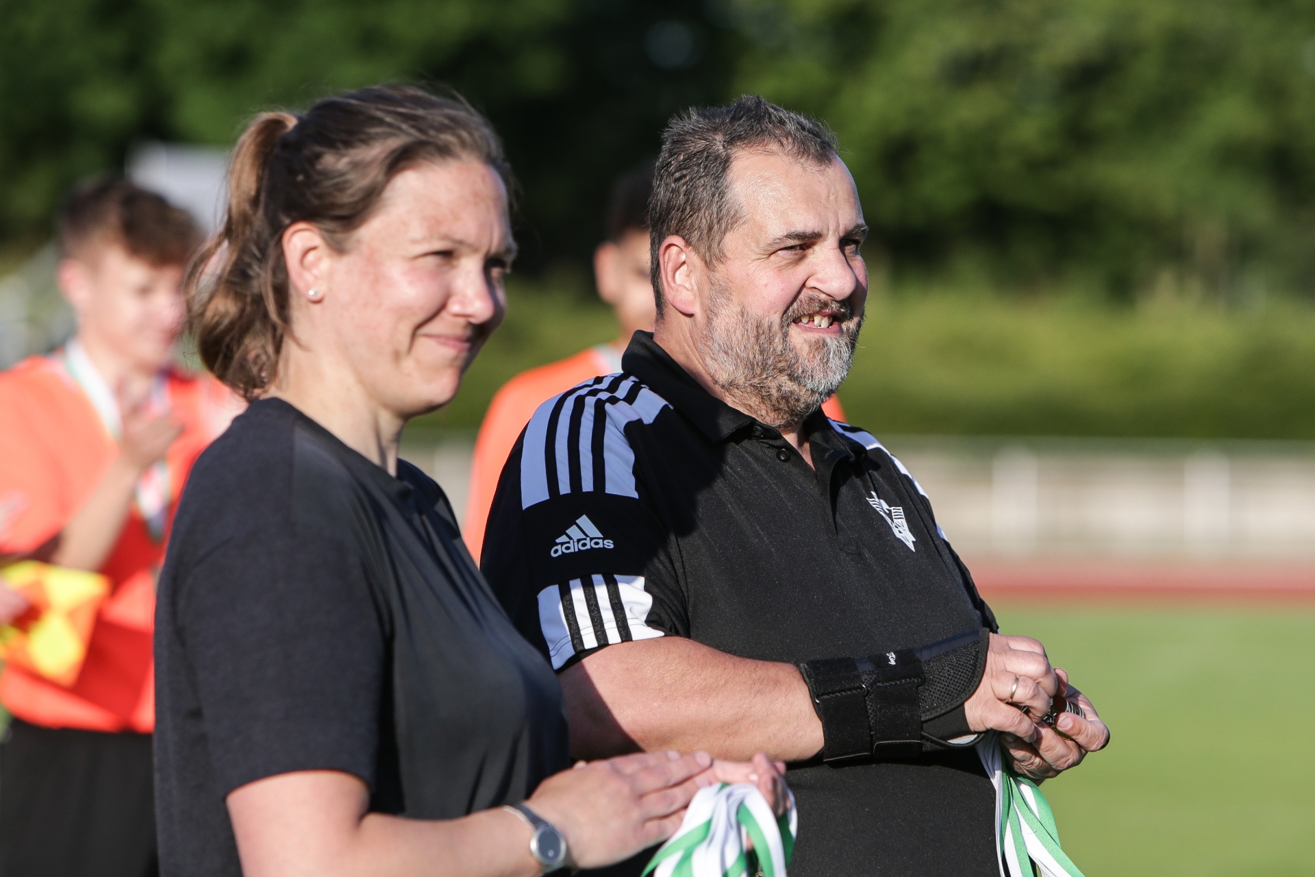 [{"clang_id":1,"value":"Zwei Schiedsrichter, eine Frau und ein Mann, stehen lächelnd auf einem Fußballfeld. Beide tragen schwarze Adidas-Kleidung und halten Flaggen. Im Hintergrund weitere Schiedsrichter."}]