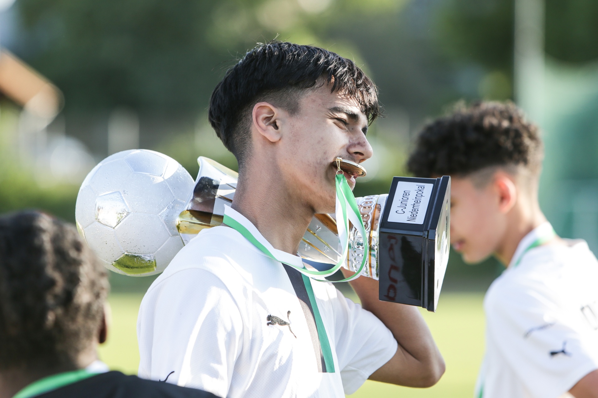 [{"clang_id":1,"value":"Junge Fußballspieler feiert Sieg. Er hält Pokal und Fußball in den Händen, jubelt mit offenem Mund. Hinter ihm weitere Spieler in Teamkleidung."}]