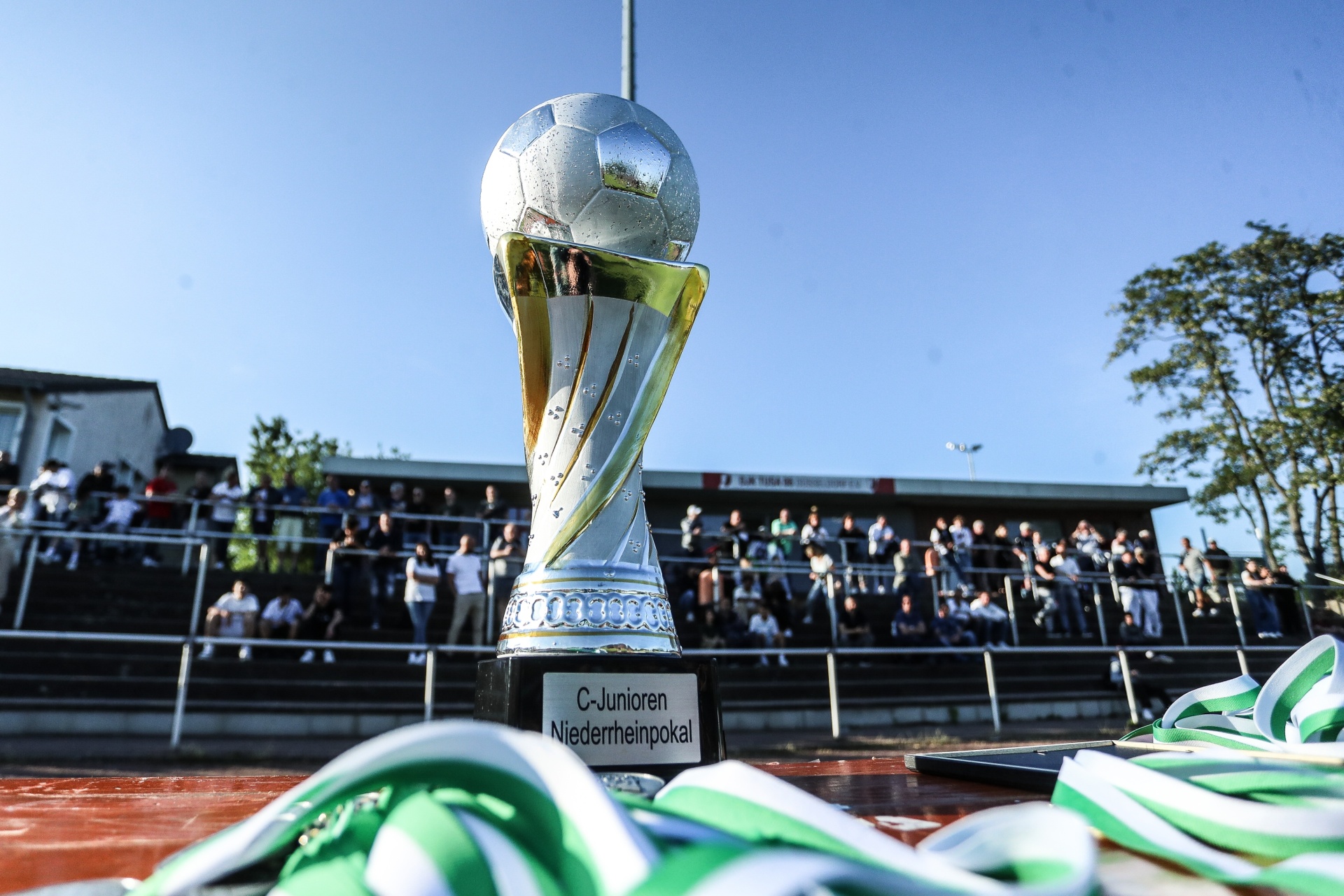 [{"clang_id":1,"value":"Fußballtrophäe im Vordergrund, darunter grün-weiße Sportbekleidung. Im Hintergrund eine Tribüne mit Zuschauern und Fußballfeld."}]