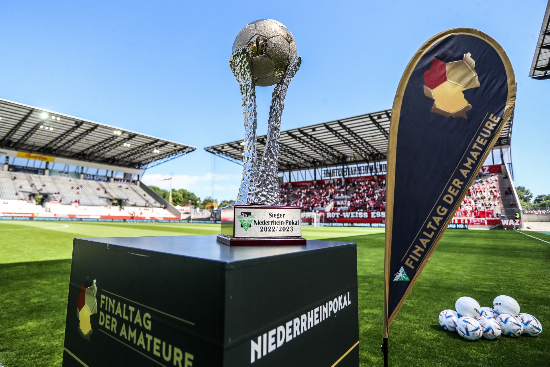 [{"clang_id":1,"value":"Fußballtrophäe auf Podest vor Stadion. Im Hintergrund Zuschauerränge und Banner \"Finaltag der Amateure\" sowie Fußbälle."}]