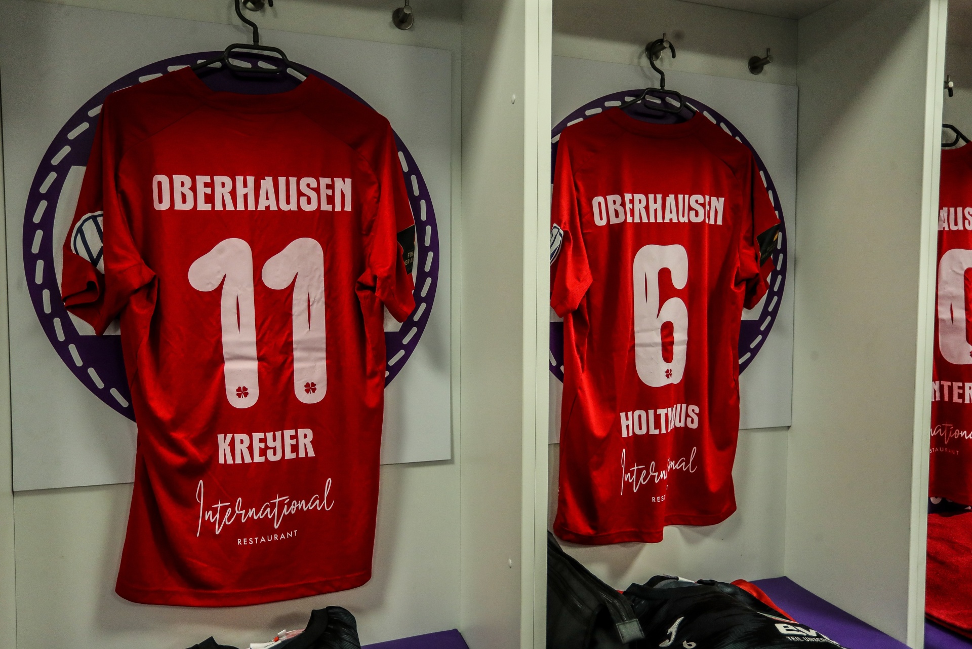 [{"clang_id":1,"value":"Zwei rote Fußballtrikots mit weißer Schrift hängen in einem dunklen Schrank. Trikot links mit Nummer 11, Kreyer, rechts mit Nummer 6, Holtus. Sponsor \"International Restaurant\" ist zu sehen."}]