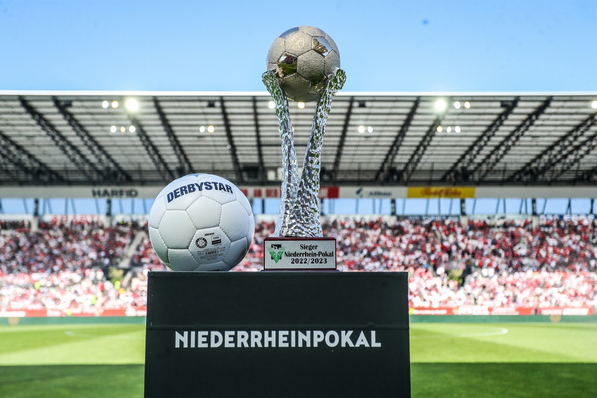 [{"clang_id":1,"value":"Fußballtrophäe \"Niederrheinpokal\" mit Fußball auf Sockel vor Stadionkulisse mit Zuschauern."}]